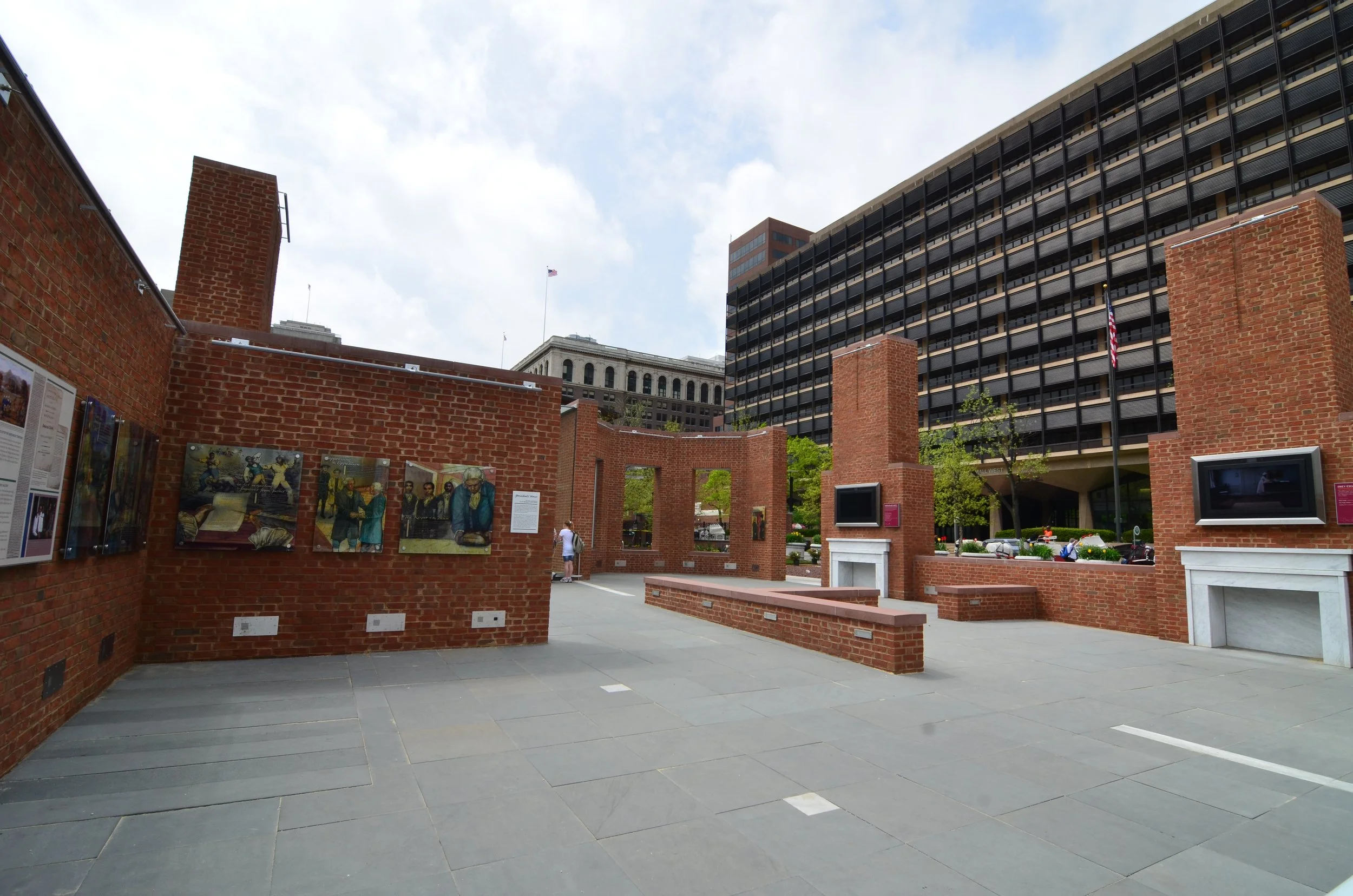 Outdoor art exhibit with brick walls, paintings, and digital screens, with city buildings and blue sky in the background.