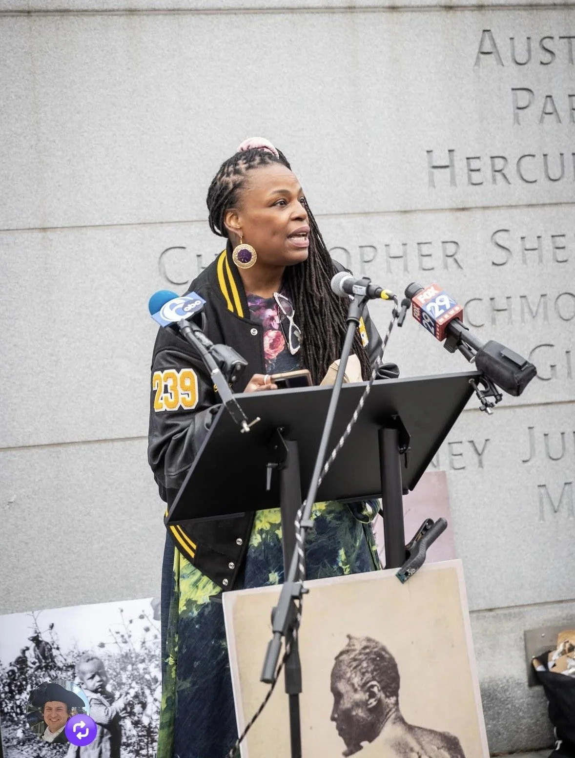 Woman speaking at a podium outdoors, with multiple microphones, wearing a black varsity jacket with yellow patches, earrings, and braids, with a grey stone wall in the background that has engraved text.