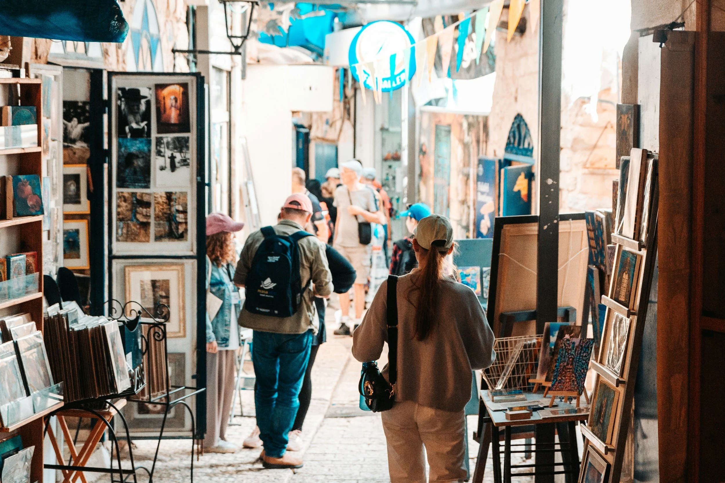People shopping at an outdoor art market with paintings displayed on boards and shelves, under a covered alleyway with rustic brick walls and hanging decorations.