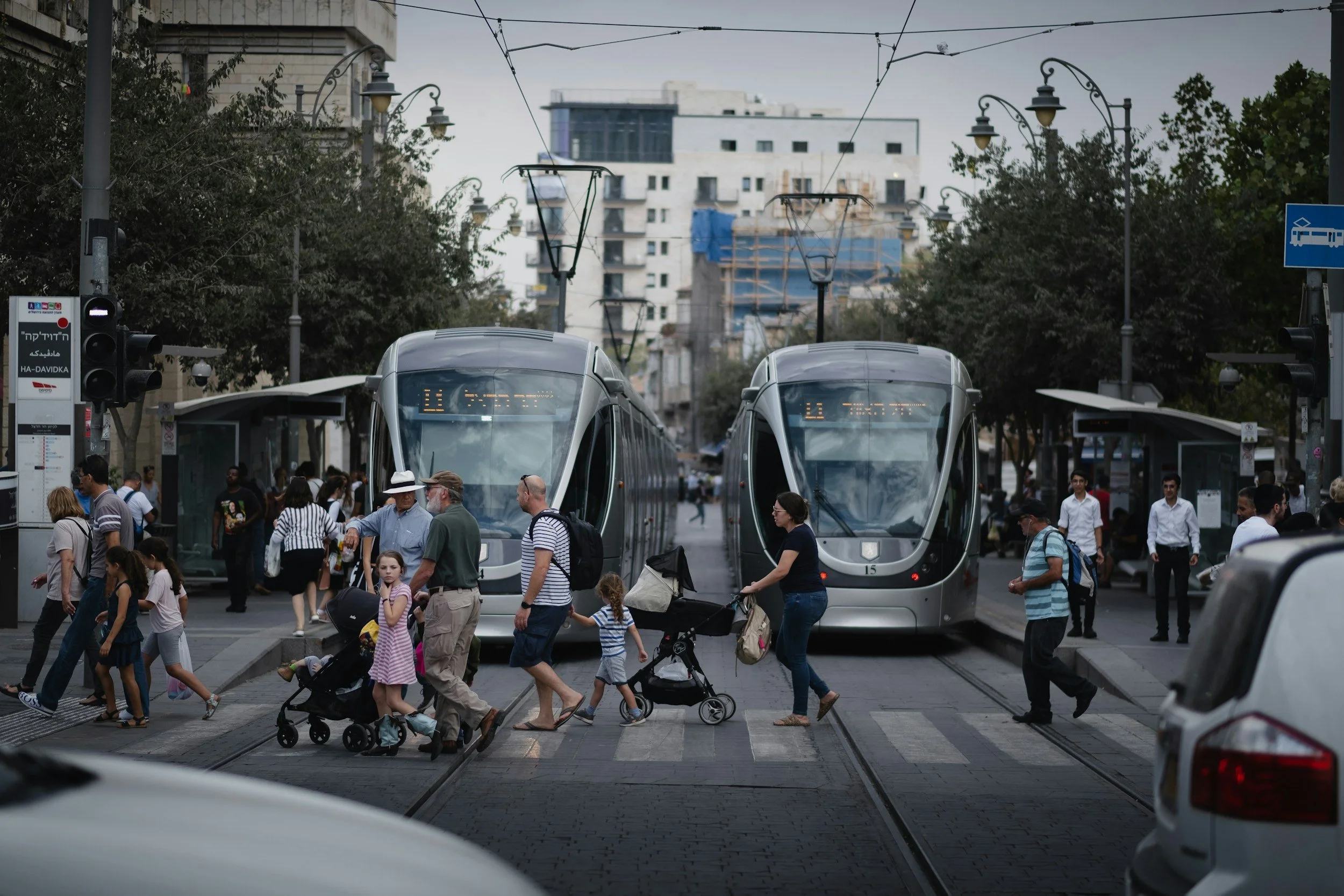 People crossing a city street with two modern trams stopped at the station, surrounded by pedestrians and urban buildings.