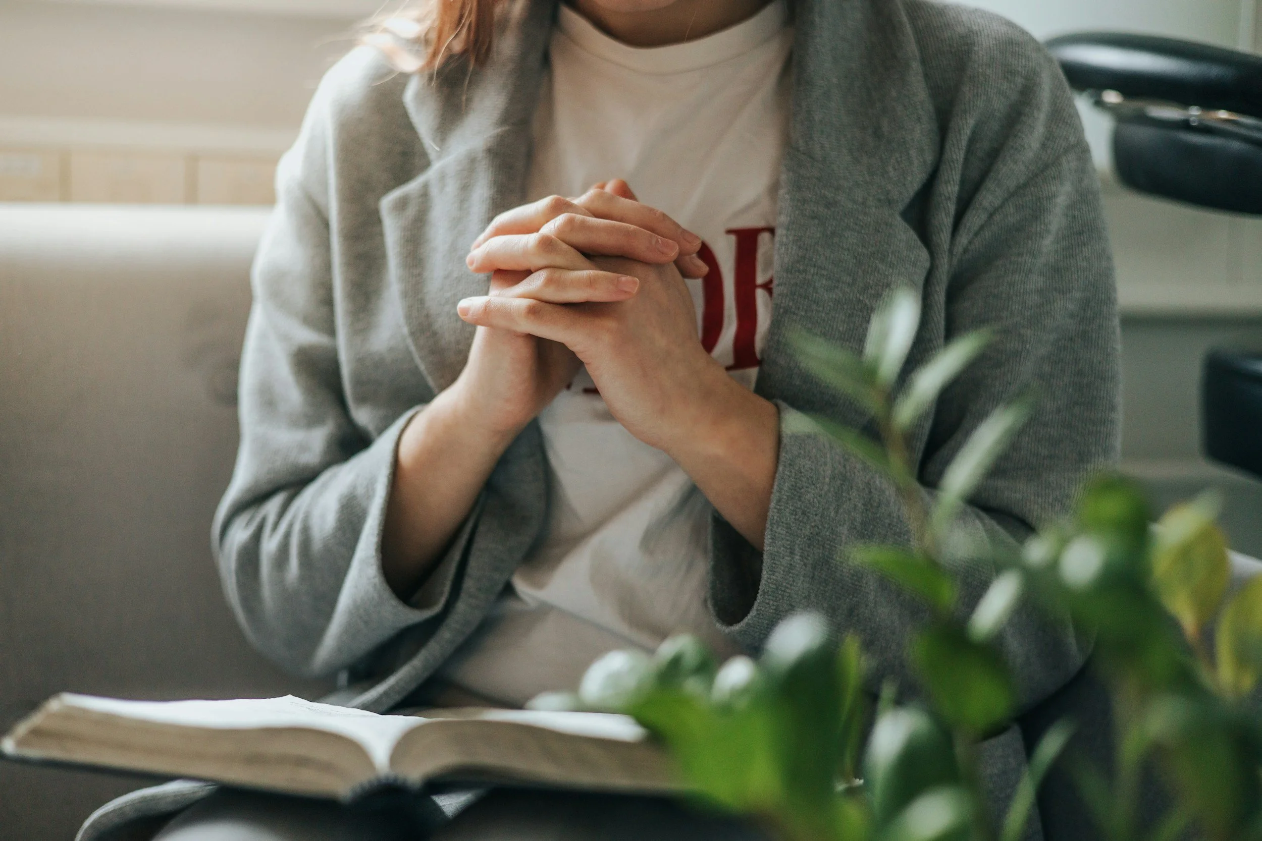 Person sitting with hands clasped over a Bible, wearing a grey blazer and white t-shirt, near some green plants.