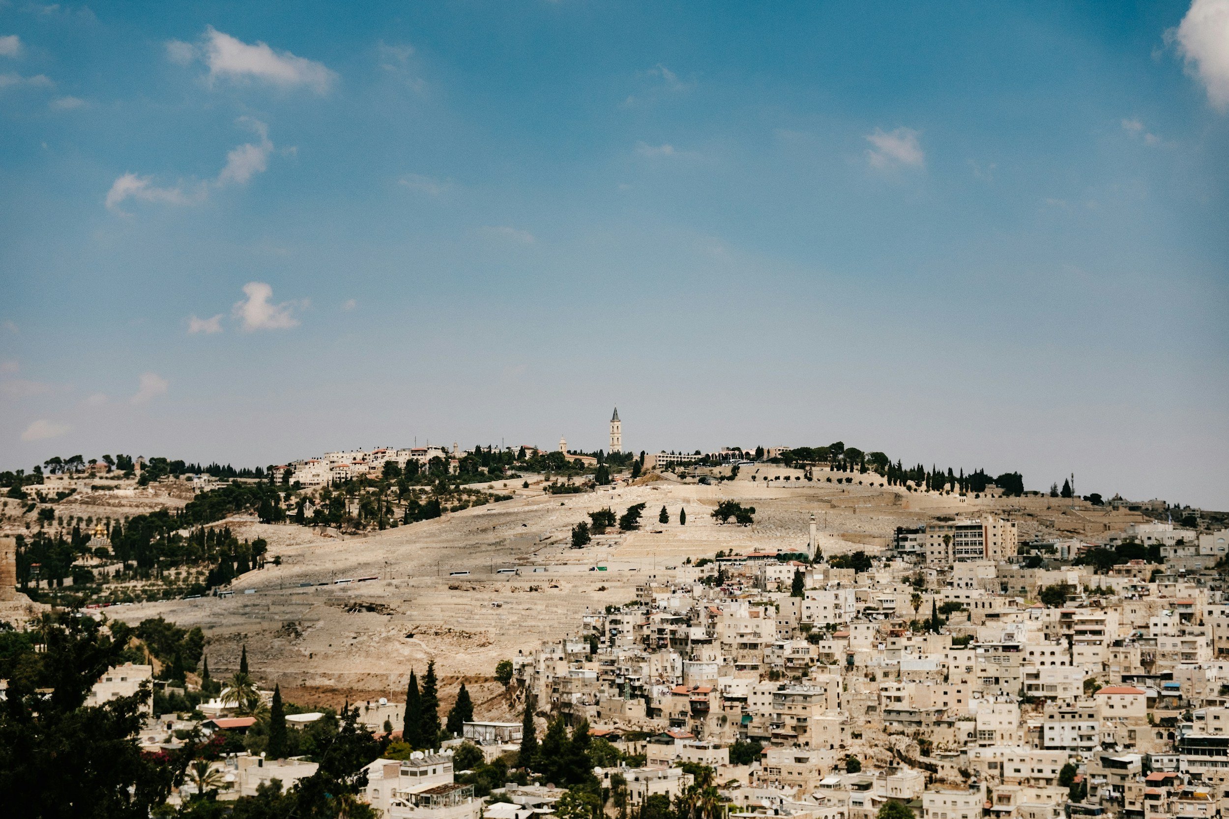 A cityscape of a hillside town with numerous white buildings, trees, and a prominent church tower, under a blue sky with scattered clouds.