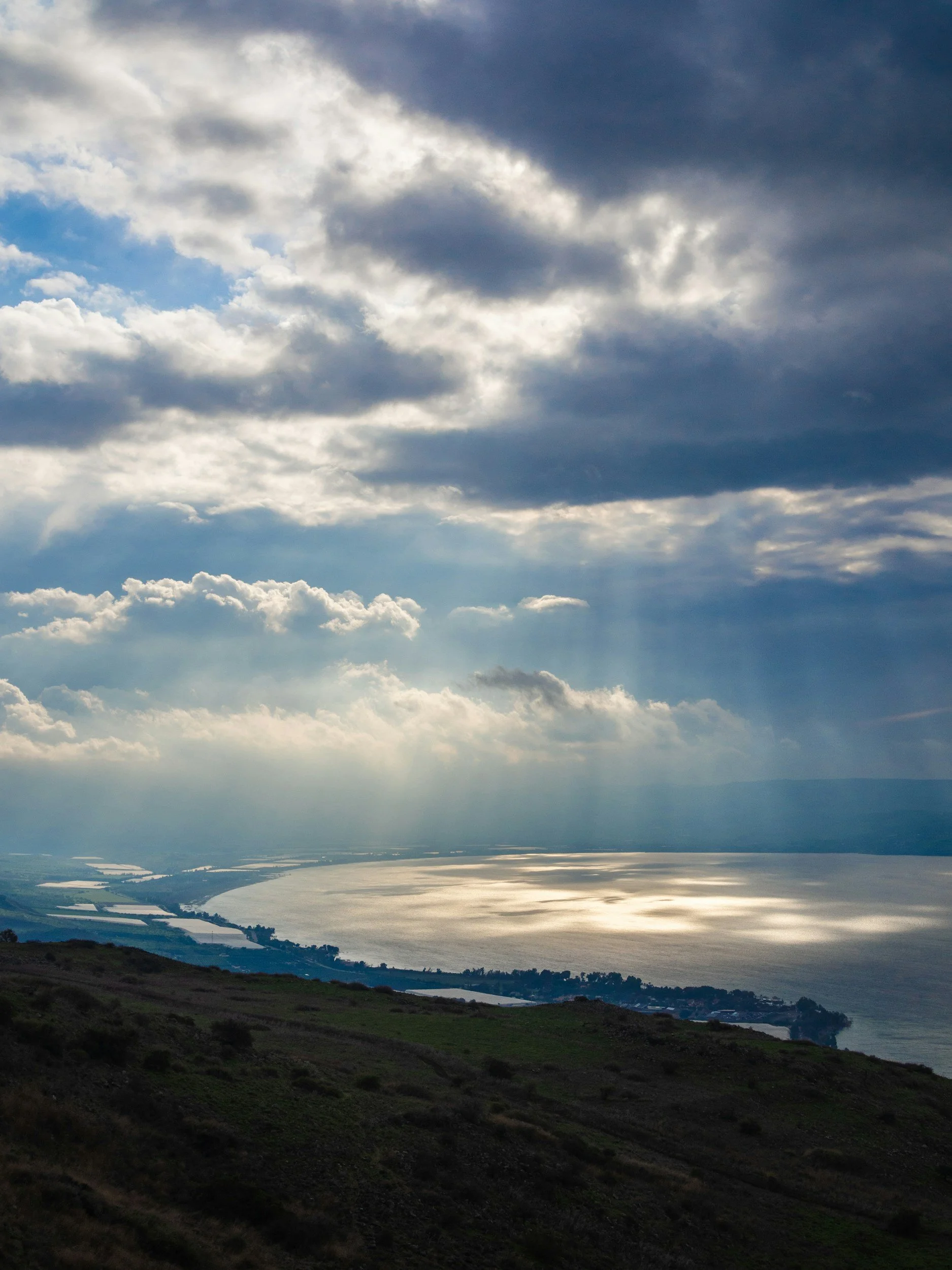 A scenic view of a lake or river with sunlight reflecting off the water, under a cloudy sky with some sun rays breaking through.