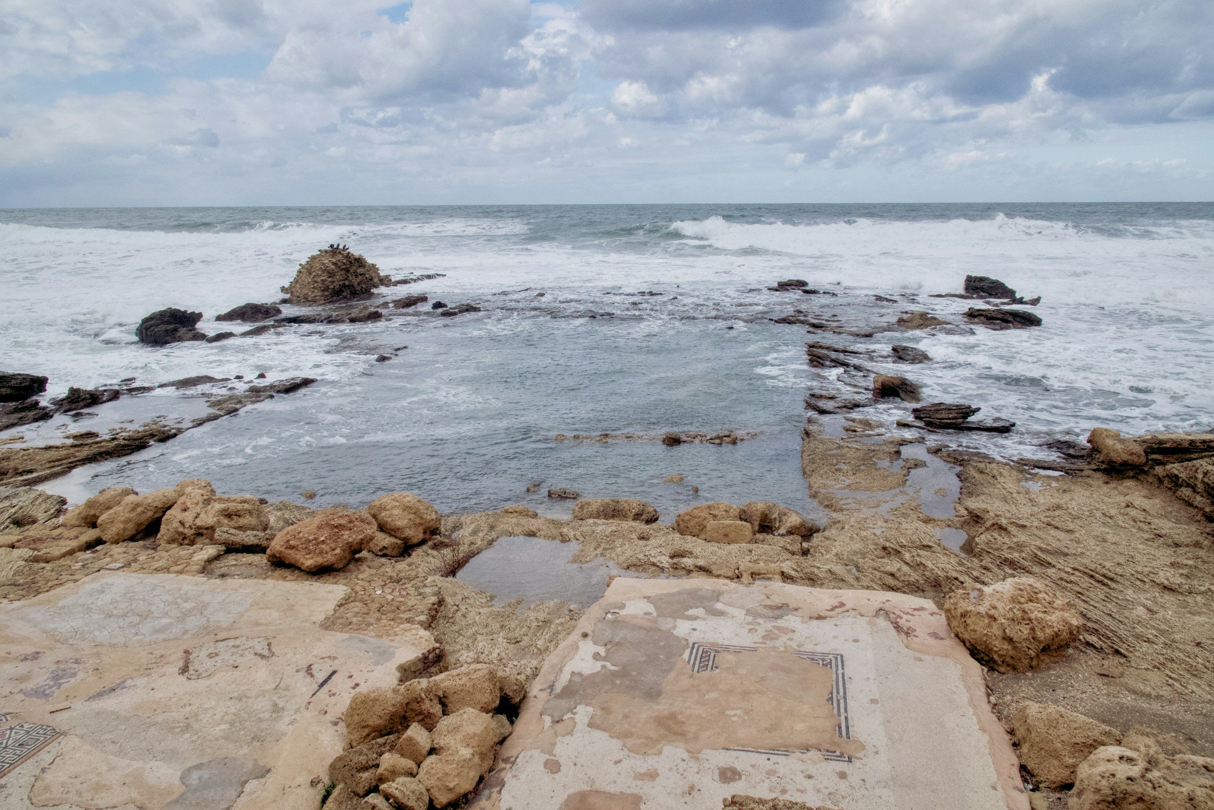 A rocky shoreline with ocean waves crashing against rocks under a partly cloudy sky.