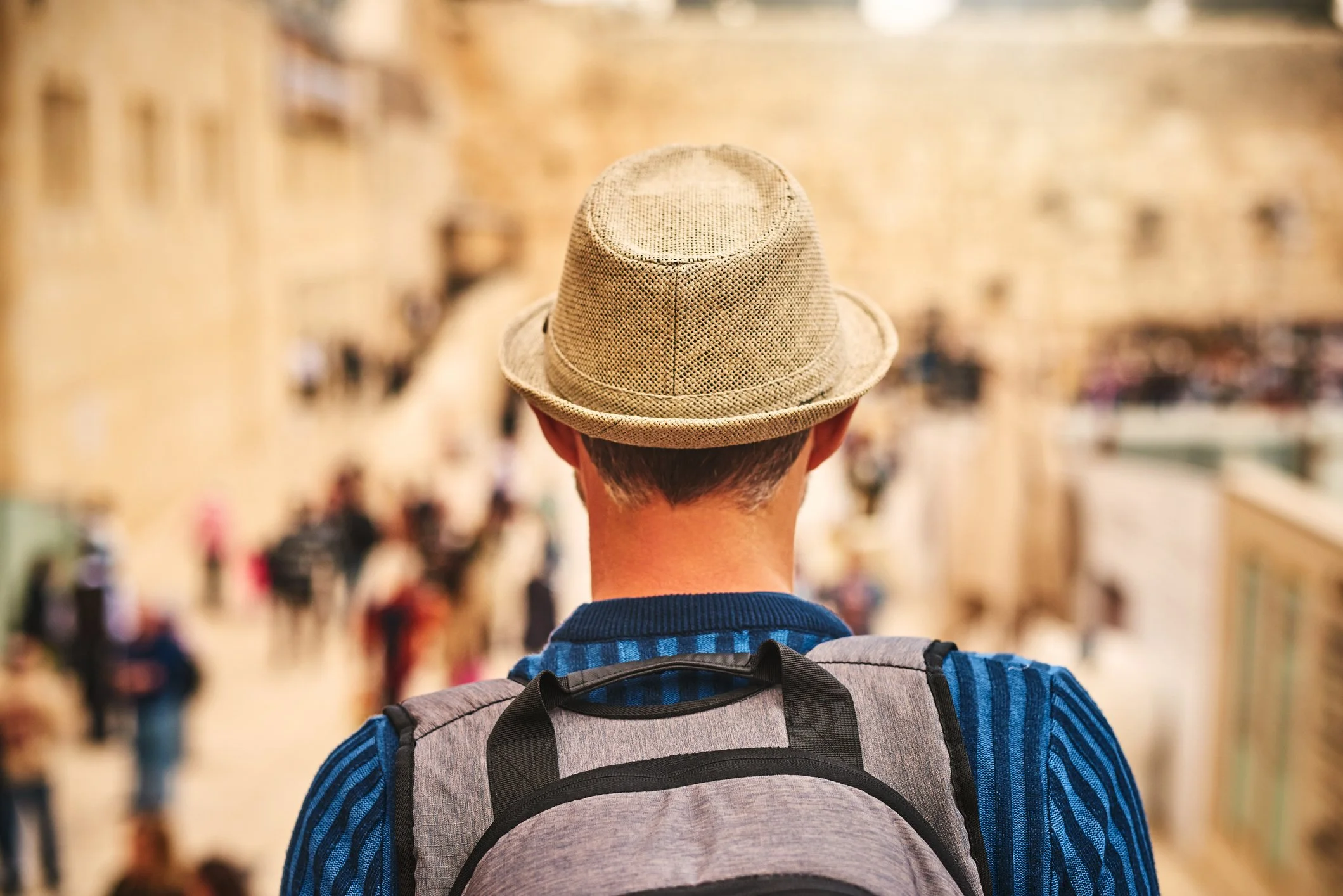A man wearing a straw fedora hat and a blue striped shirt with a gray backpack is viewed from behind, in a crowded outdoor market or street.