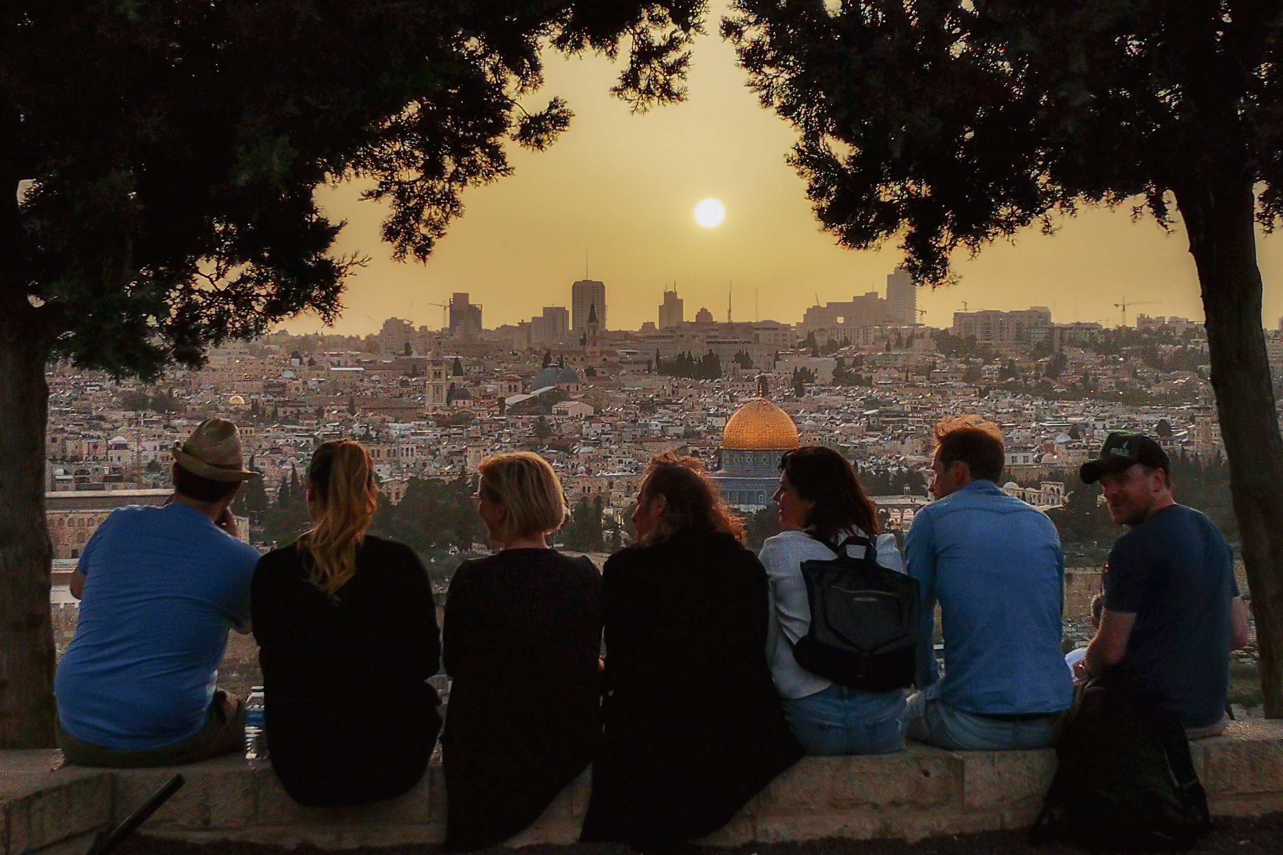 Group of people sitting on a stone wall overlooking a cityscape at sunset, with the golden Dome of the Rock prominent in the background, framed by trees.