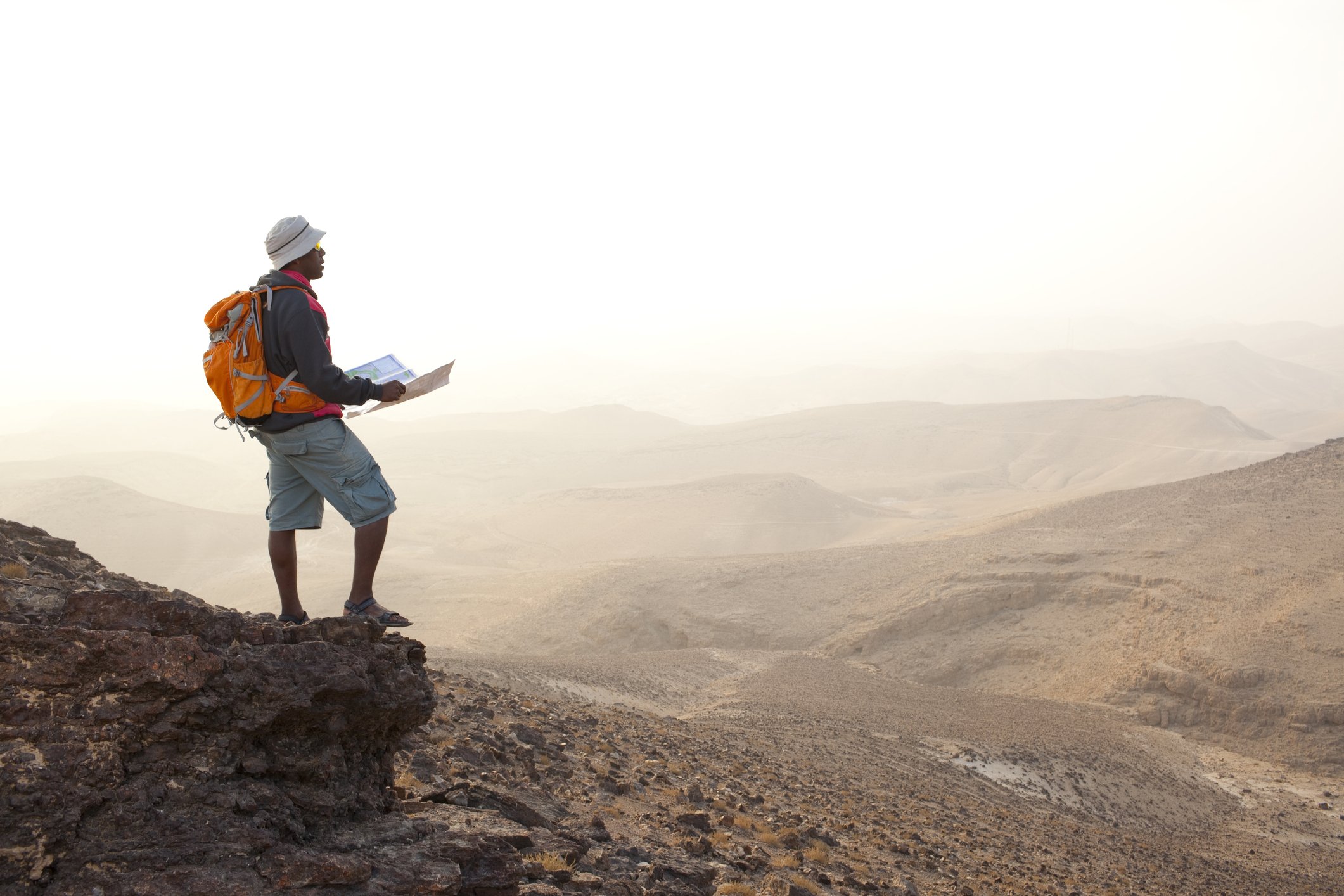 A man standing on a rocky ledge in a desert landscape, holding a map and looking into the distance, with an orange backpack and a hat.