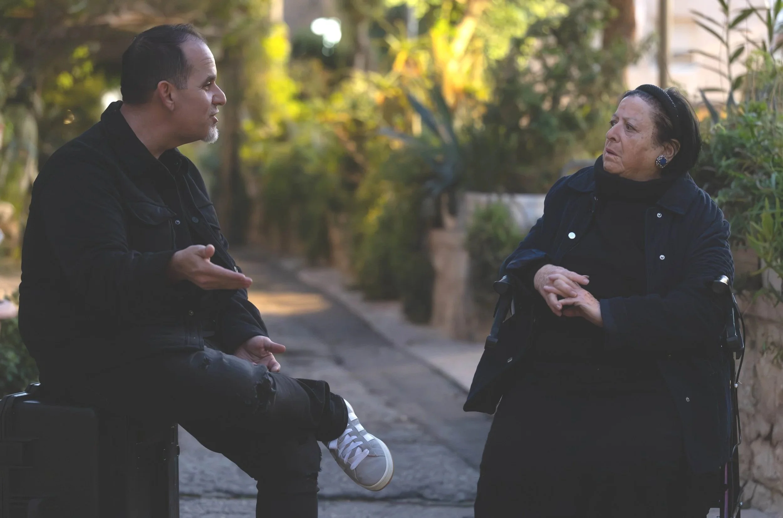 A man and an elderly woman in conversation outdoors on a sidewalk, with trees and plants in the background, during daylight.