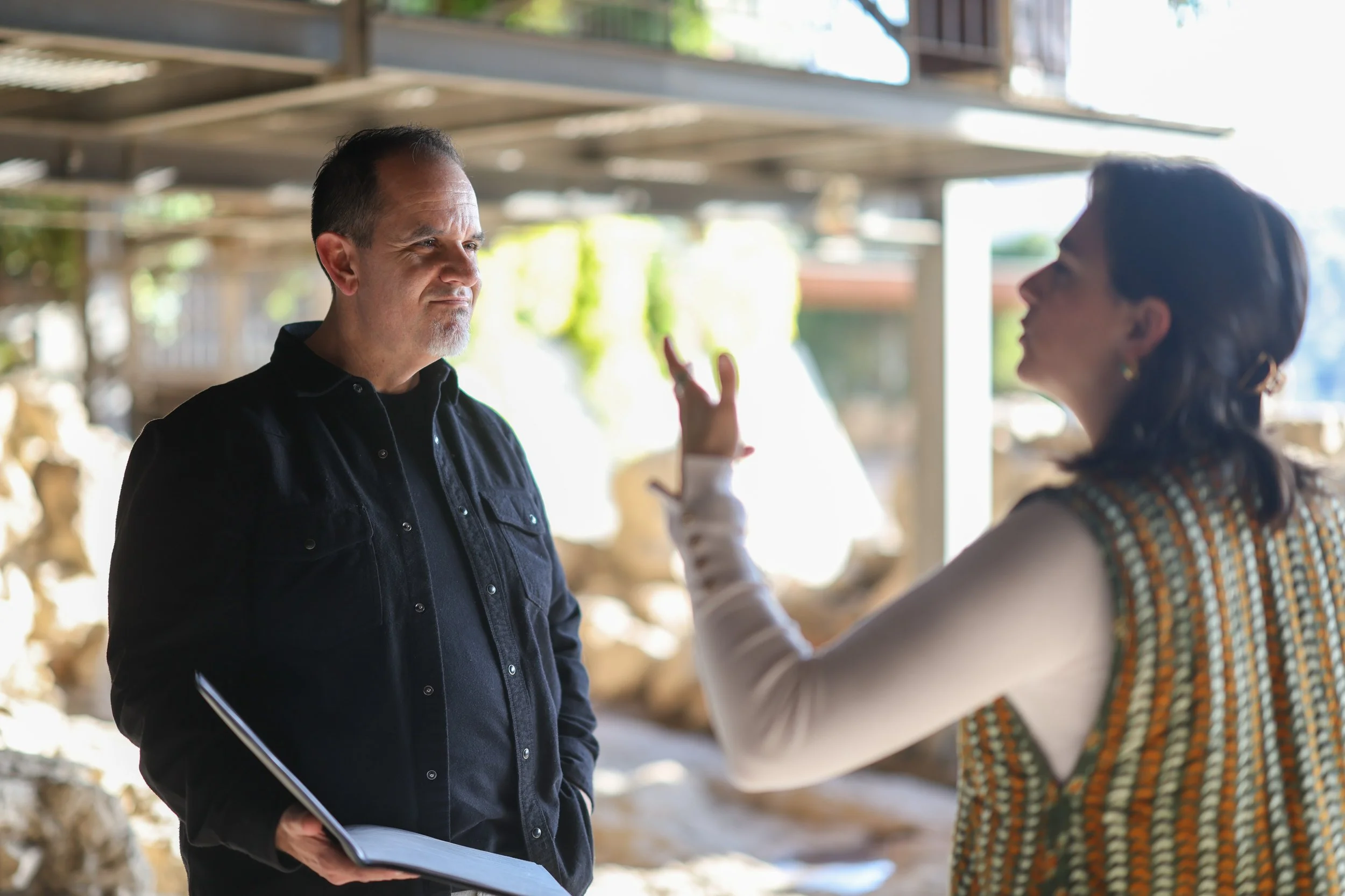 A woman is gesturing with her hand while talking to a man who is holding a folder, in an outdoor or semi-outdoor setting with a blurred background of wood logs and structures.