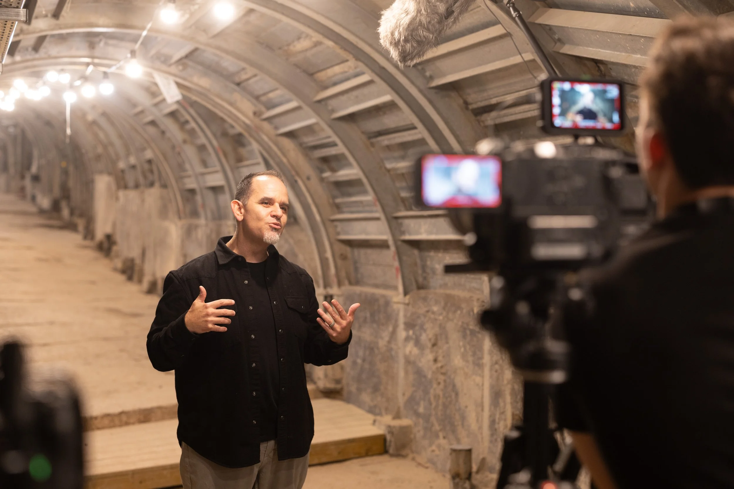 Man giving a presentation or interview inside a tunnel with arched walls, being recorded by a camera operator.