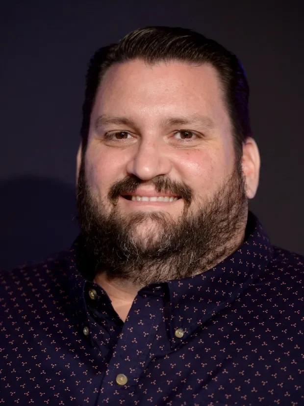 Headshot of a man with dark hair, beard, and a friendly smile, wearing a dark patterned shirt against a dark background.