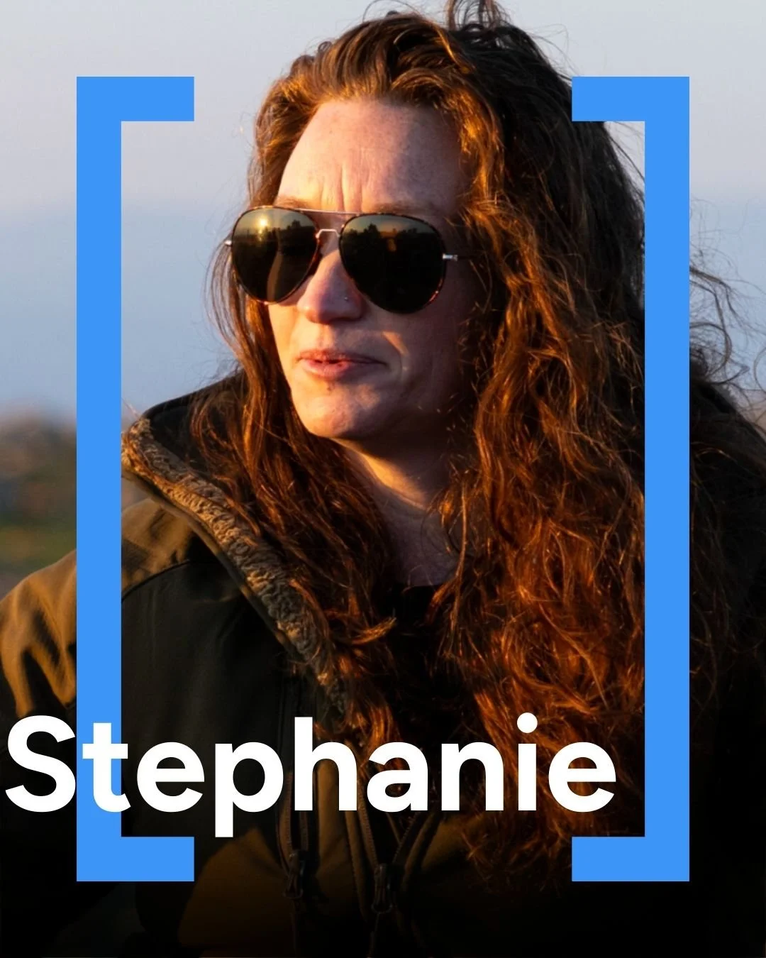 Portrait of a woman with curly brown hair wearing sunglasses, a black jacket, looking to the side during sunset.