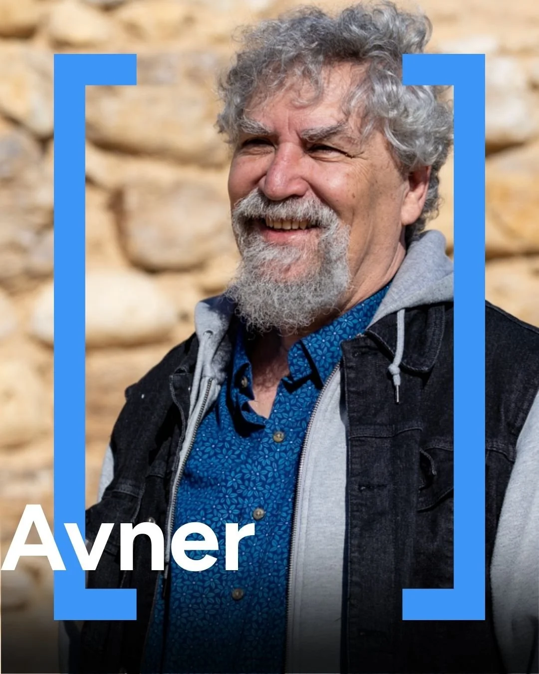 A smiling older man with gray curly hair and a beard, wearing a blue patterned shirt and a black vest, standing outdoors with a stone wall in the background.
