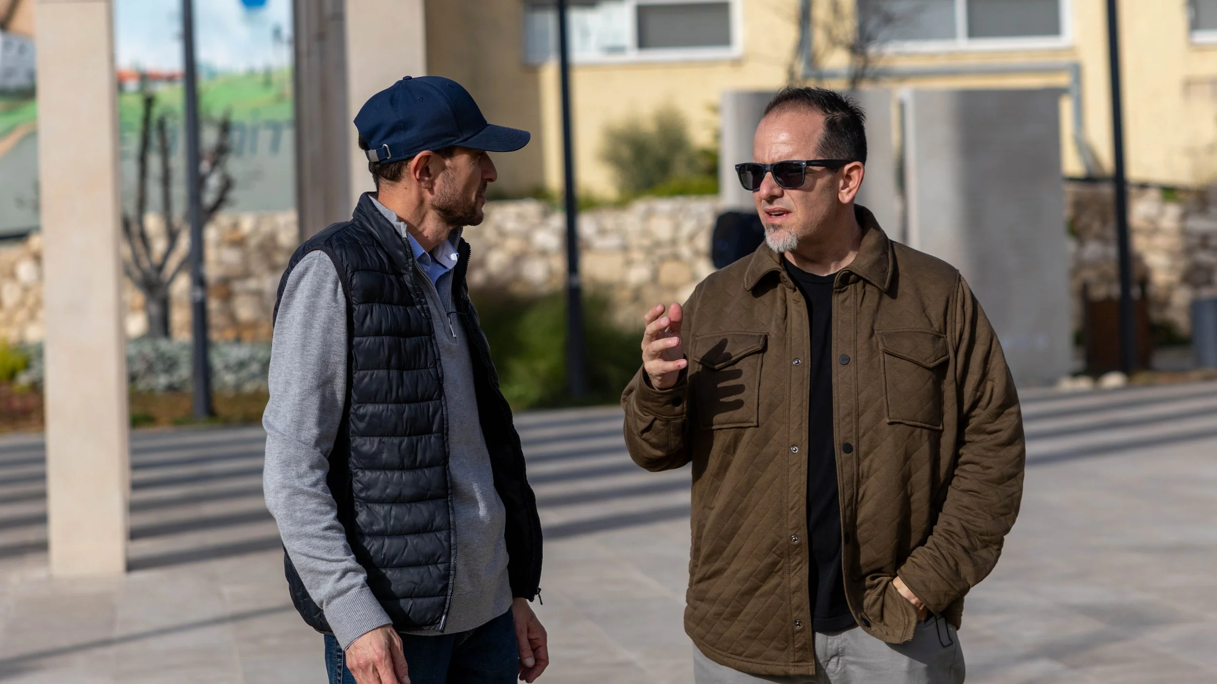 Two men are engaged in a conversation outdoors on a city sidewalk. One man is wearing a navy cap, gray sweater, and black puffer vest, while the other is wearing sunglasses, a brown jacket, and a black shirt. They are standing near a building with a stone wall and trees in the background.