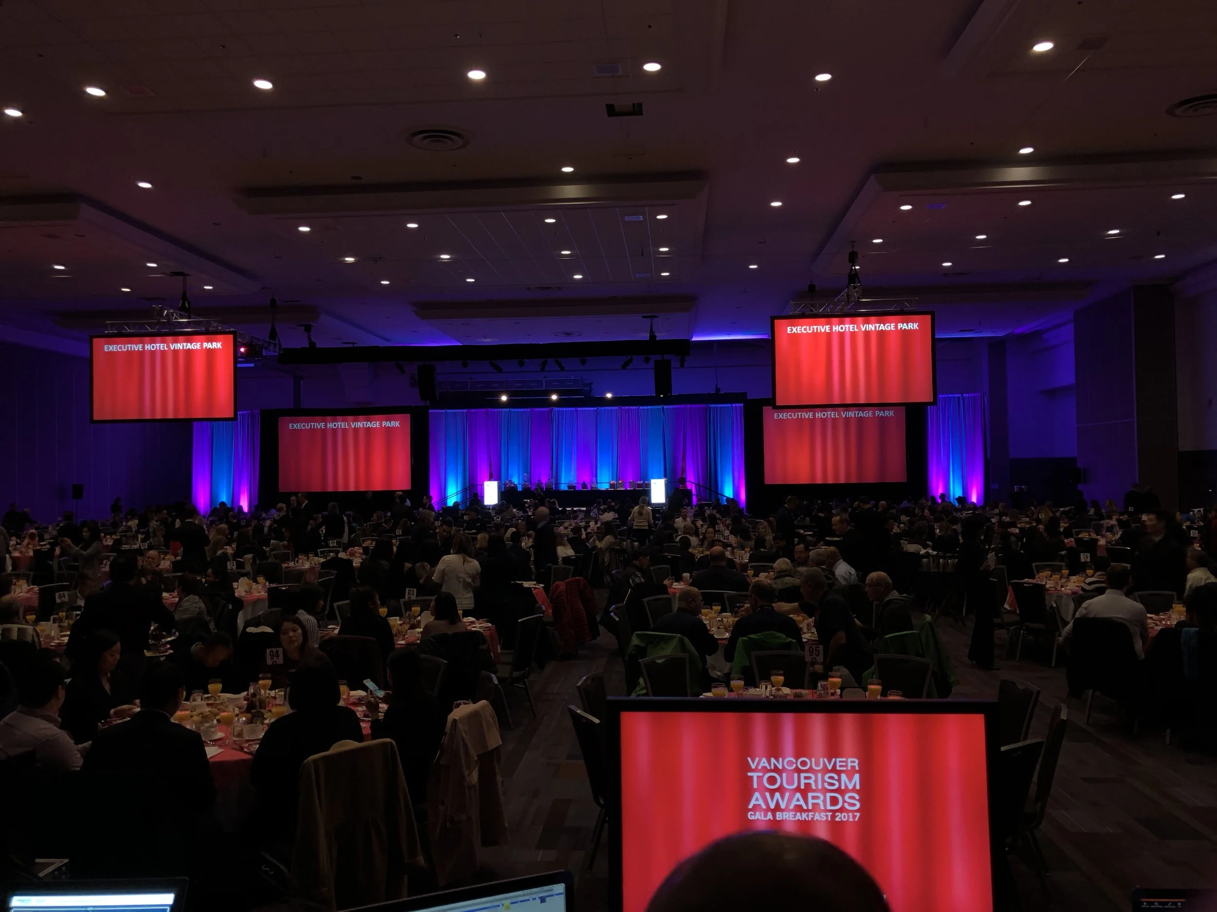 A large banquet hall filled with round tables set for the Vancouver Tourism Awards Gala Breakfast 2017. The stage has multiple screens displaying red backgrounds with white text, including 'Executive Hotel Vintage Park' and 'Vancouver Tourism Awards Gala Breakfast 2017'. The room is decorated with blue and purple lighting.