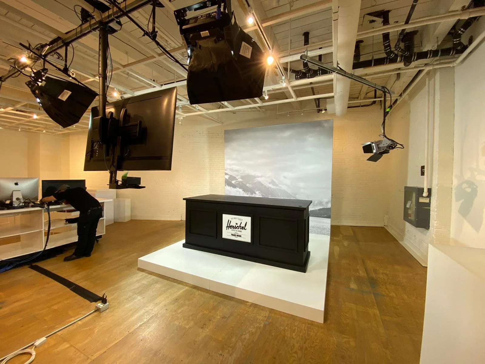 Studio with lighting equipment, a black podium with a Herschel signage, and a large backdrop of a mountain landscape.