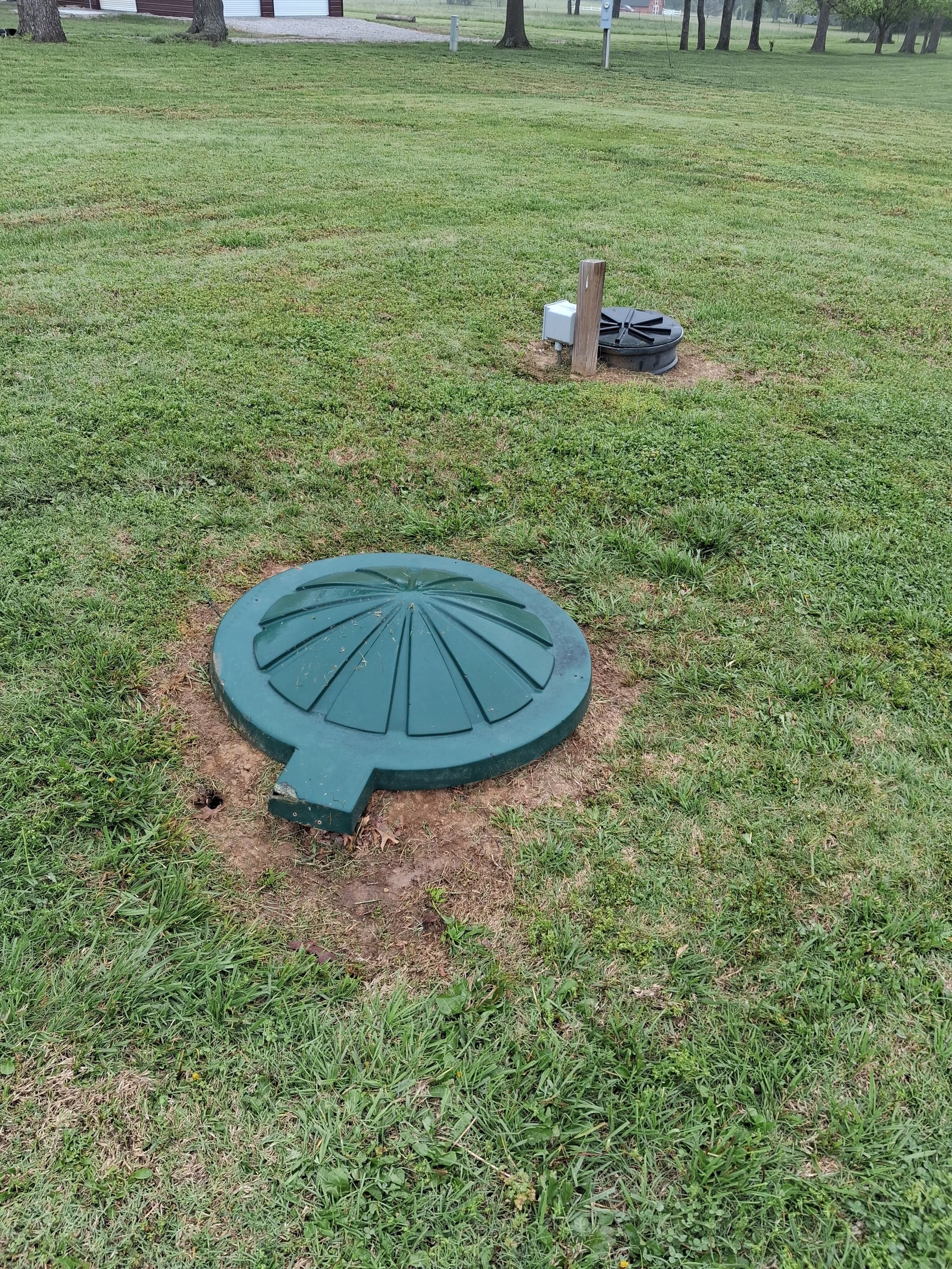 Disassembled ground cover or manhole cover with a large round green lid lying on grass, with a black base and a wooden post nearby in an open grassy field.