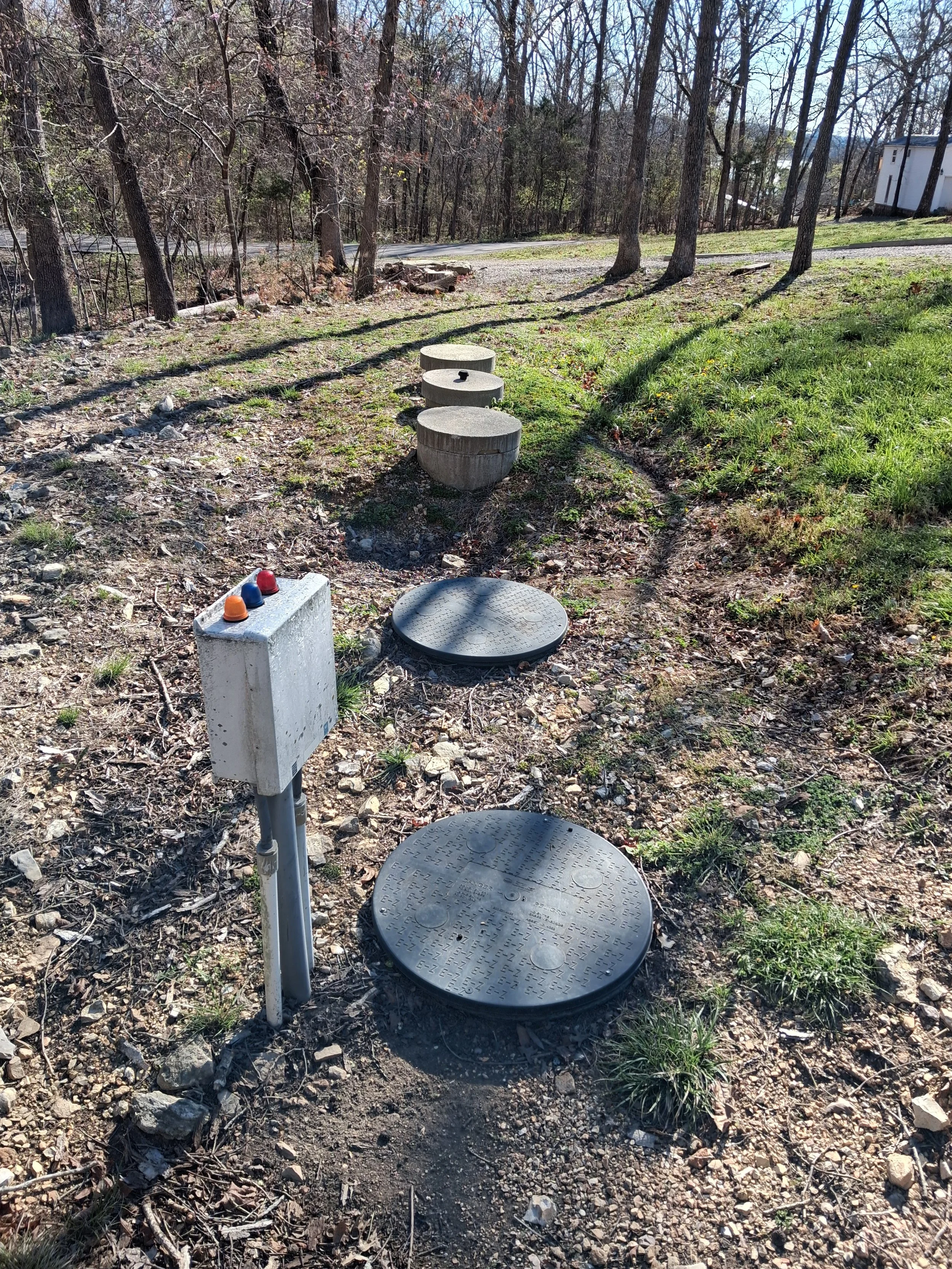 Manhole covers and utility box in a grassy outdoor area with trees and a house in the background.