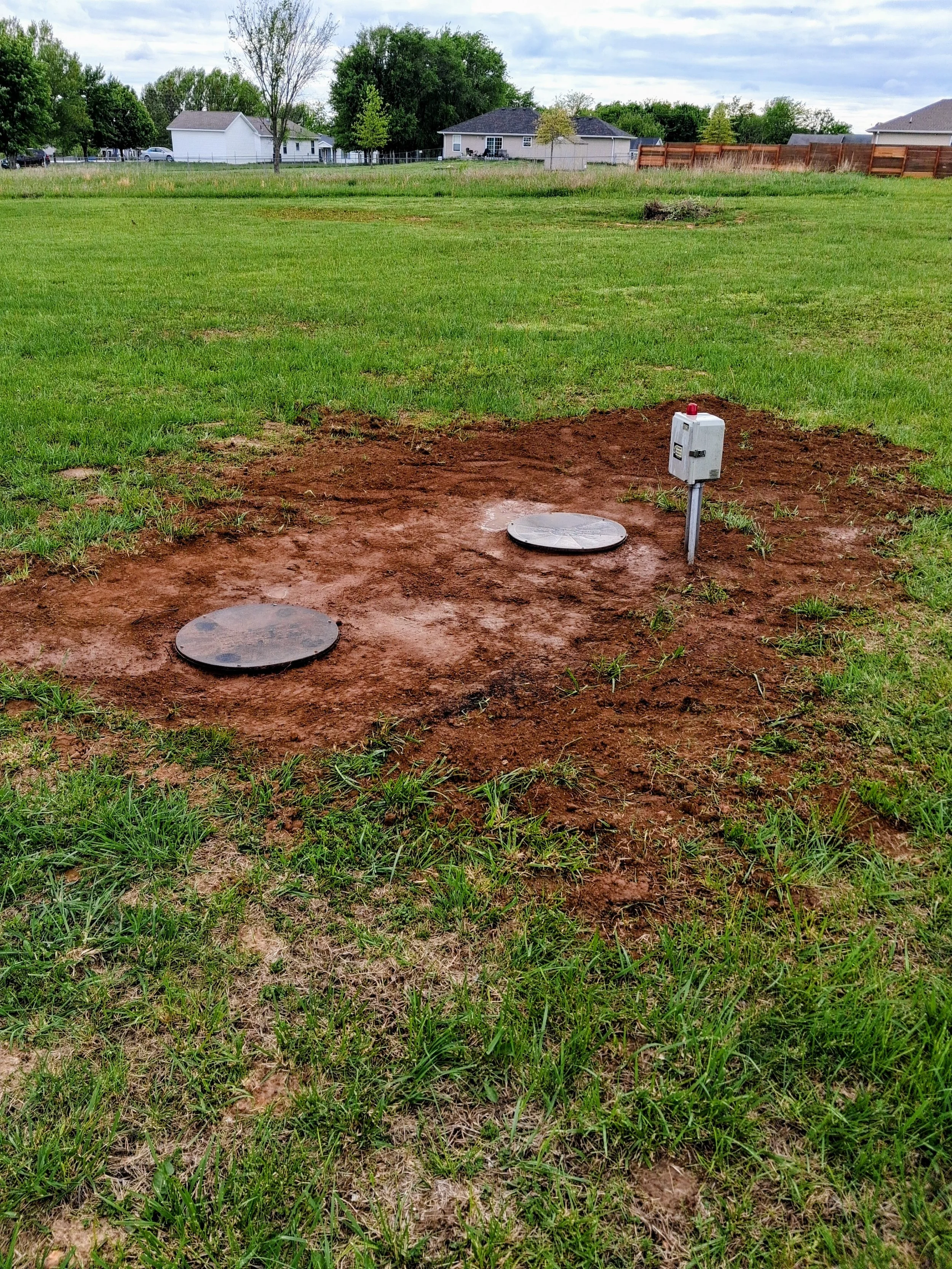 Empty patch of soil with two manhole covers and a utility box installed on a grassy field. Houses and trees are visible in the background under a cloudy sky.