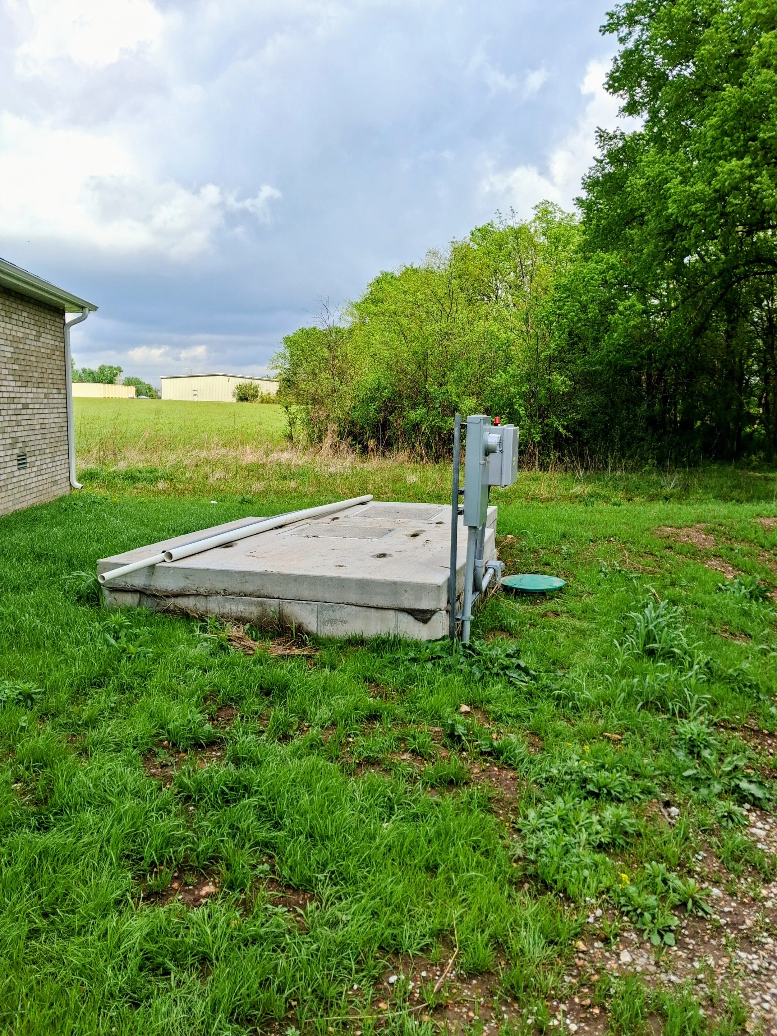 A grassy backyard with a concrete slab and white pipes, adjacent to a brick house with a white gutter, next to trees and a cloudy sky.