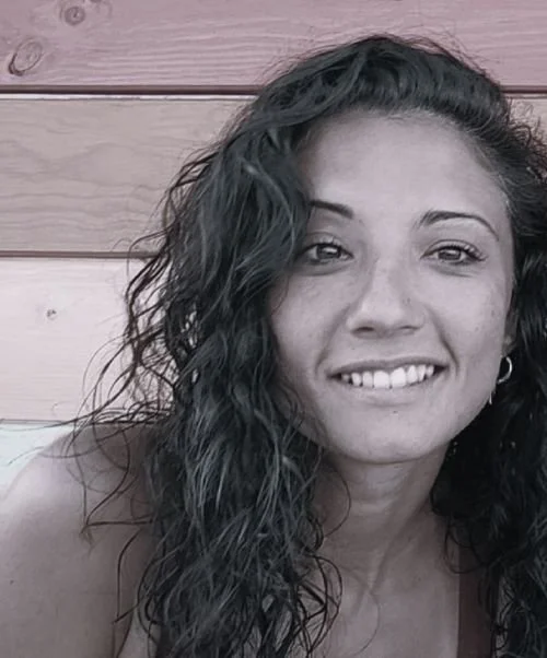 Retrato de una mujer con cabello rizado y oscuro, sonriendo y usando un pendiente, en un fondo de madera.