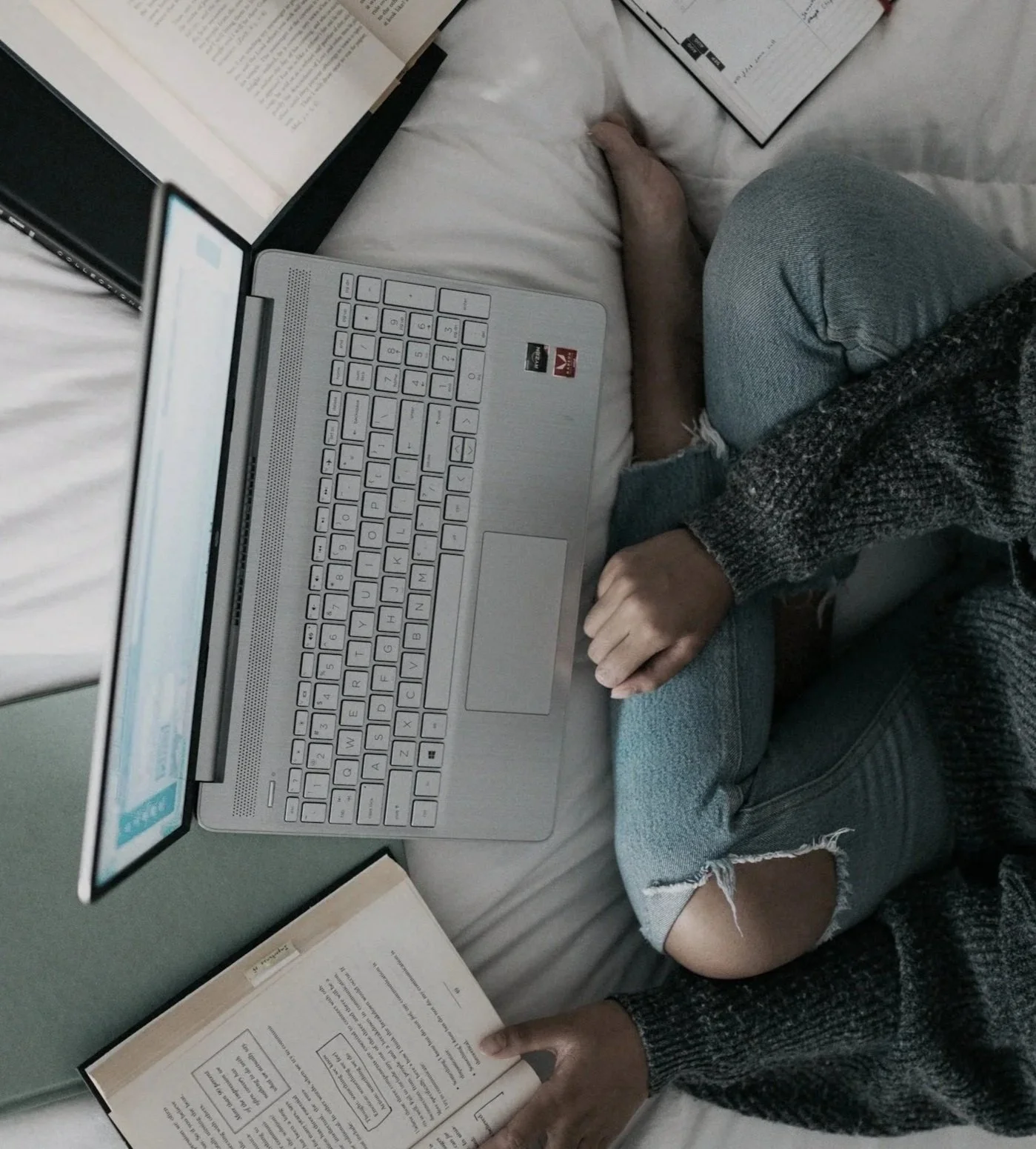Una persona estudiando en una cama, con una computadora portátil y libros abiertos, vestida con ropa casual y medias rasgadas.