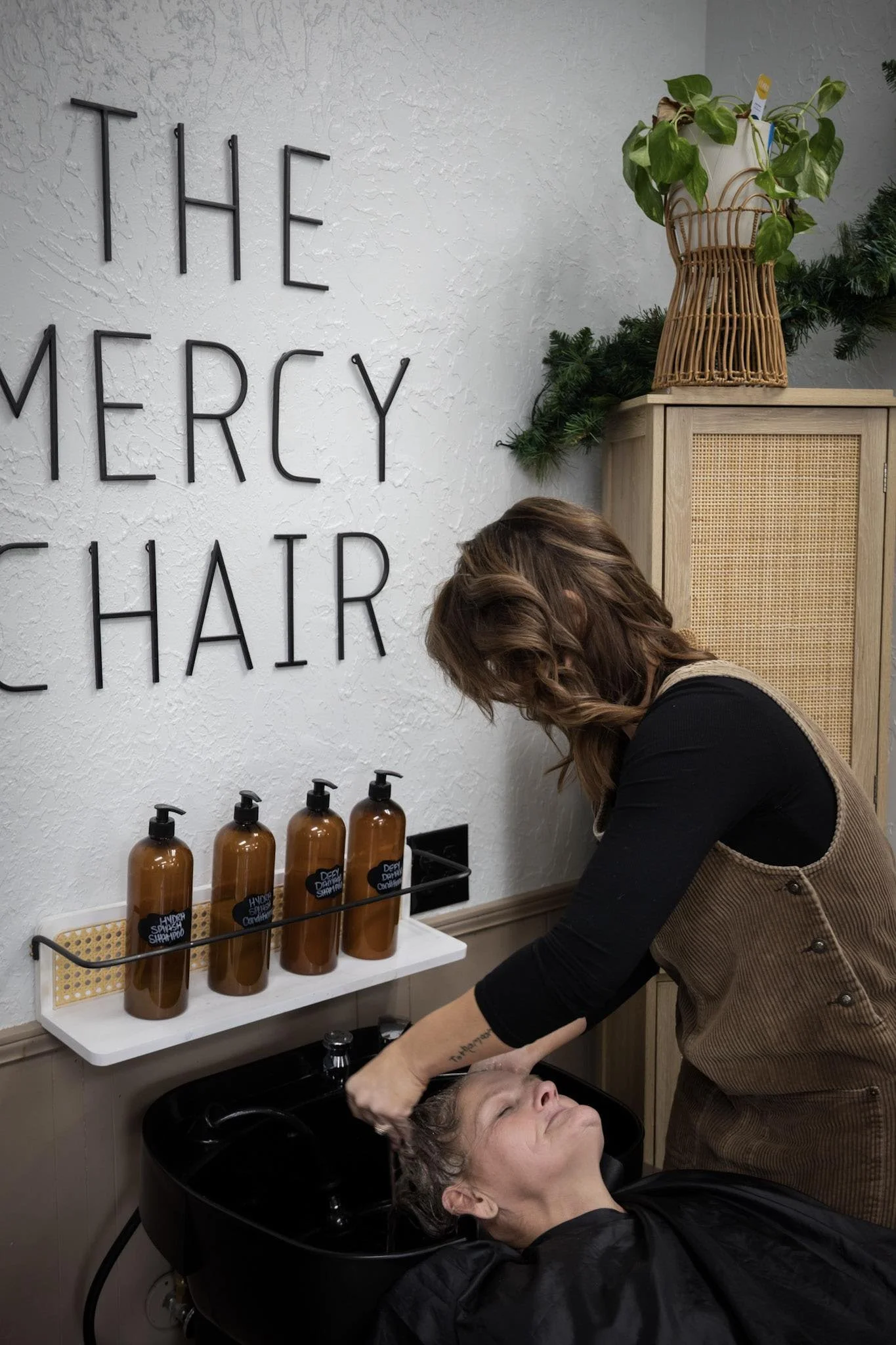 A woman getting her hair washed at a salon. The salon has a white textured wall with the words 'THE MERCY HAIR' in black letters. There are four brown shampoo bottles on a white shelf above a black sink, where the woman is leaning back with her eyes closed.