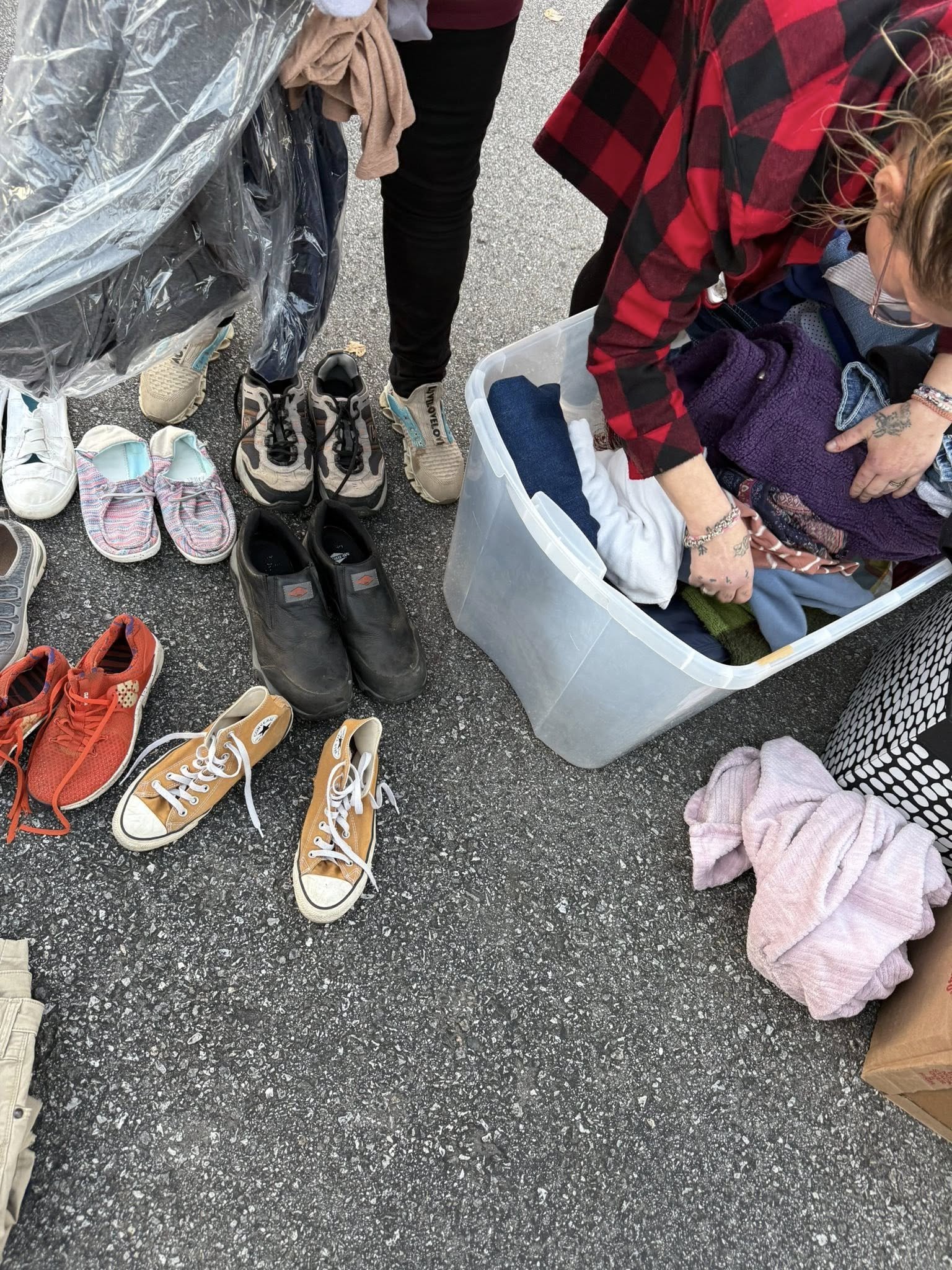 Person sorting through clothes in a large plastic bin at an outdoor flea market, with shoes arranged on the ground nearby.