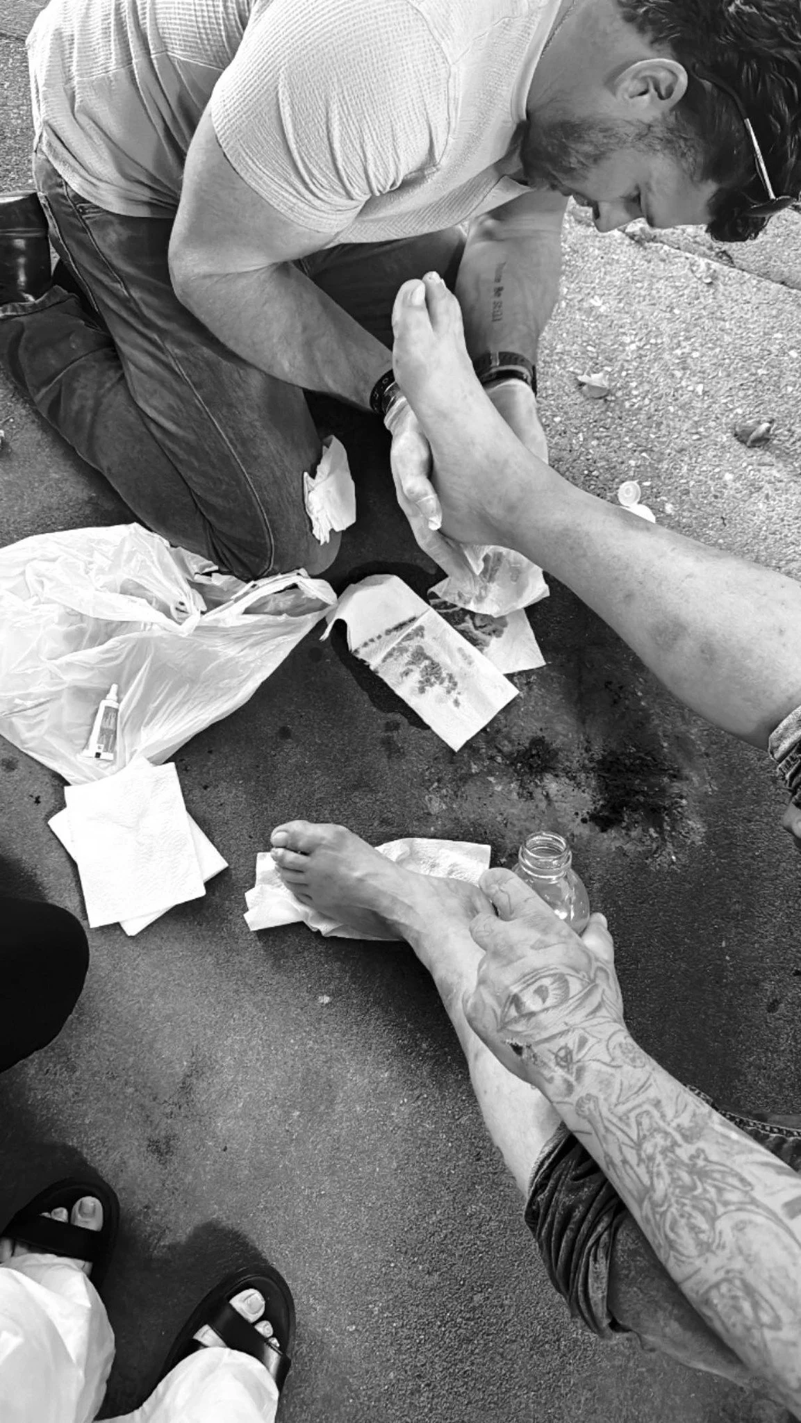 A person with tattoos on their arm holding a bottle of water and a foot with a wound, another person tending to the wound on the ground with medical supplies around.