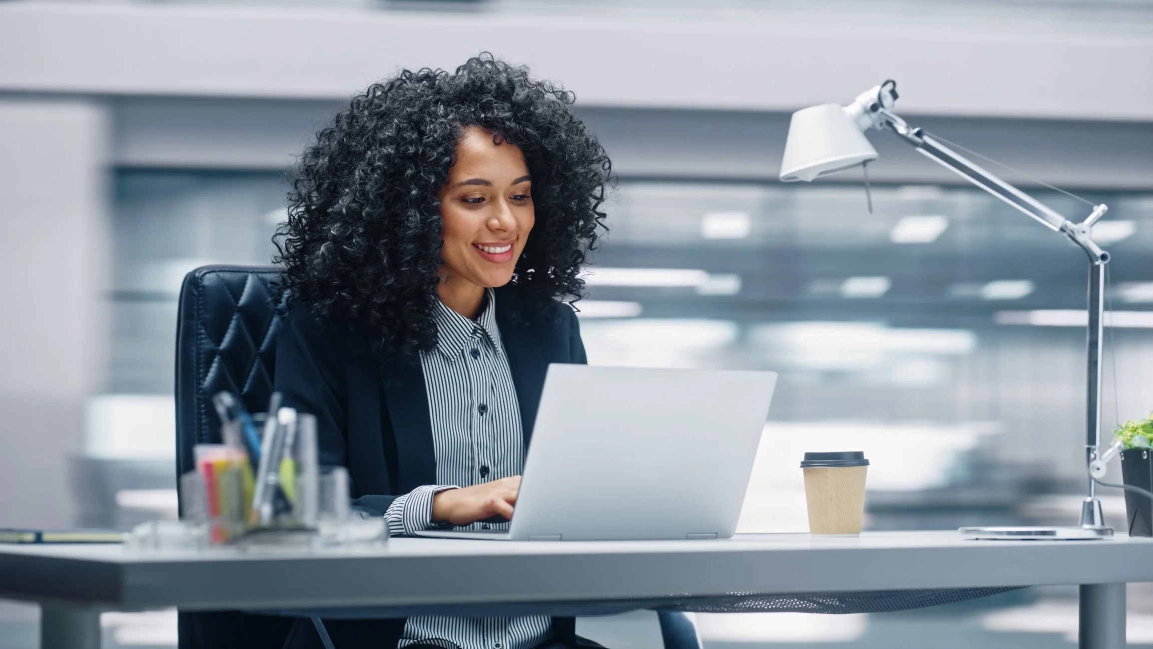 A woman with curly black hair is sitting at a desk in an office, working on a laptop, with a coffee cup, desk lamp, and office supplies around her.