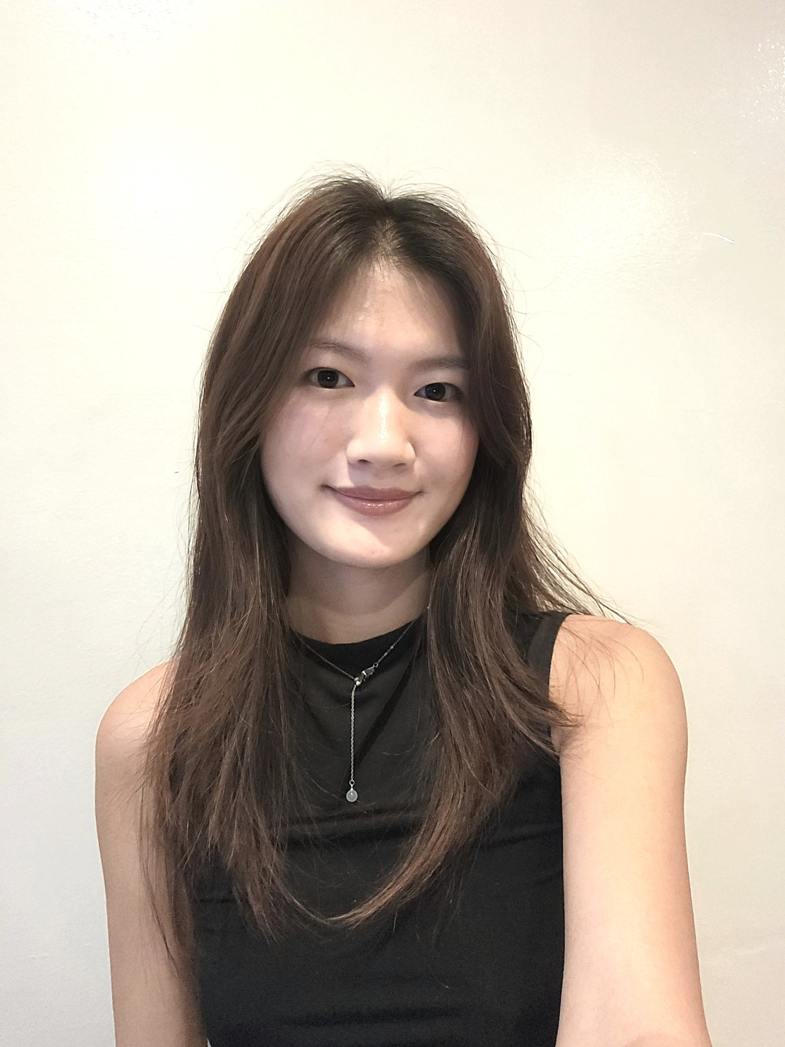A young woman with long, brown hair, wearing a black sleeveless top and a silver necklace, smiling at the camera against a plain light-colored wall.
