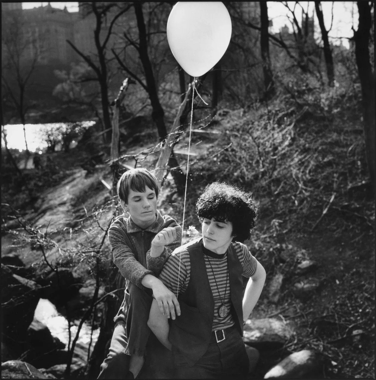 Two young men in a wooded outdoor setting, one sitting on the other's back, holding a balloon, with bare trees and a small body of water in the background.