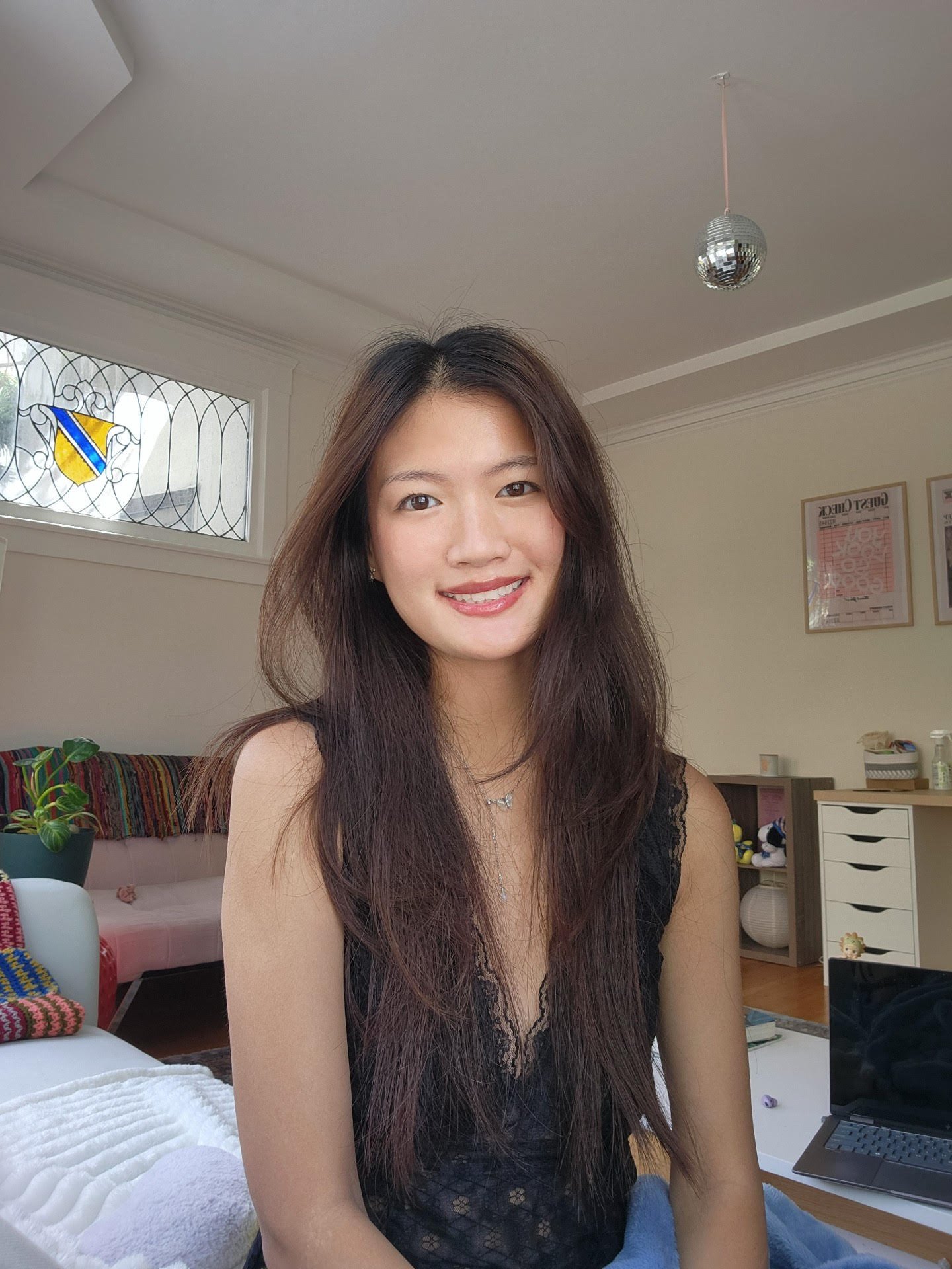A smiling woman with long dark hair sitting in a bright room, with a kitchen and furnishings in the background.