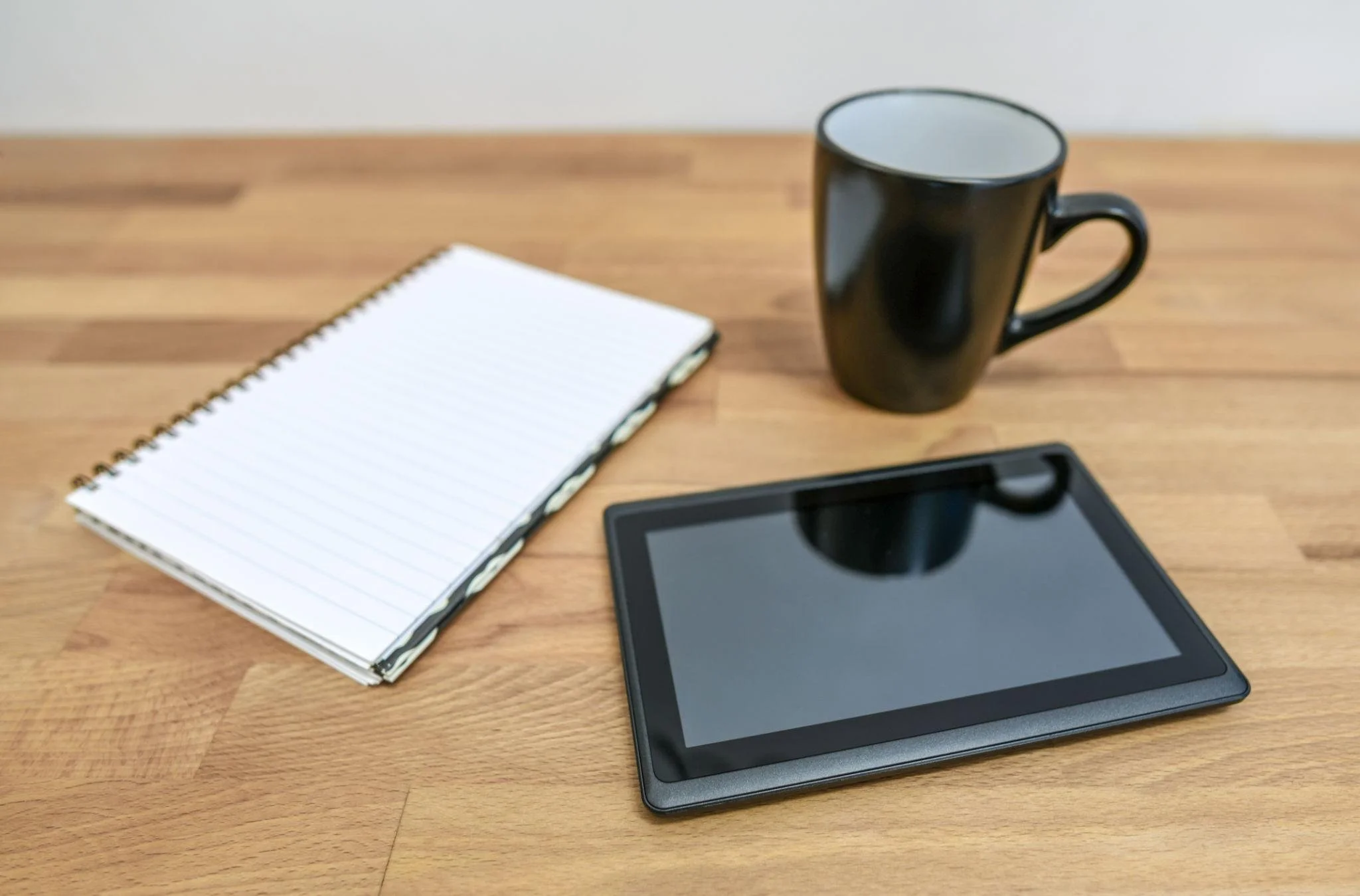 A wooden desk with a black tablet, a spiral notepad, a black coffee mug, and part of a smartphone.
