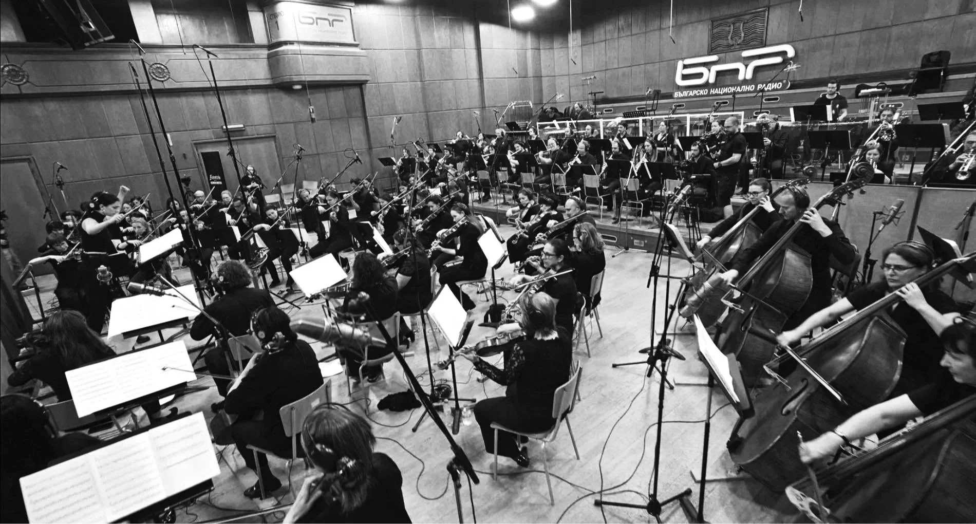 An orchestra in a recording studio with musicians playing various instruments including violins, cellos, and woodwinds, and a conductor leading them.