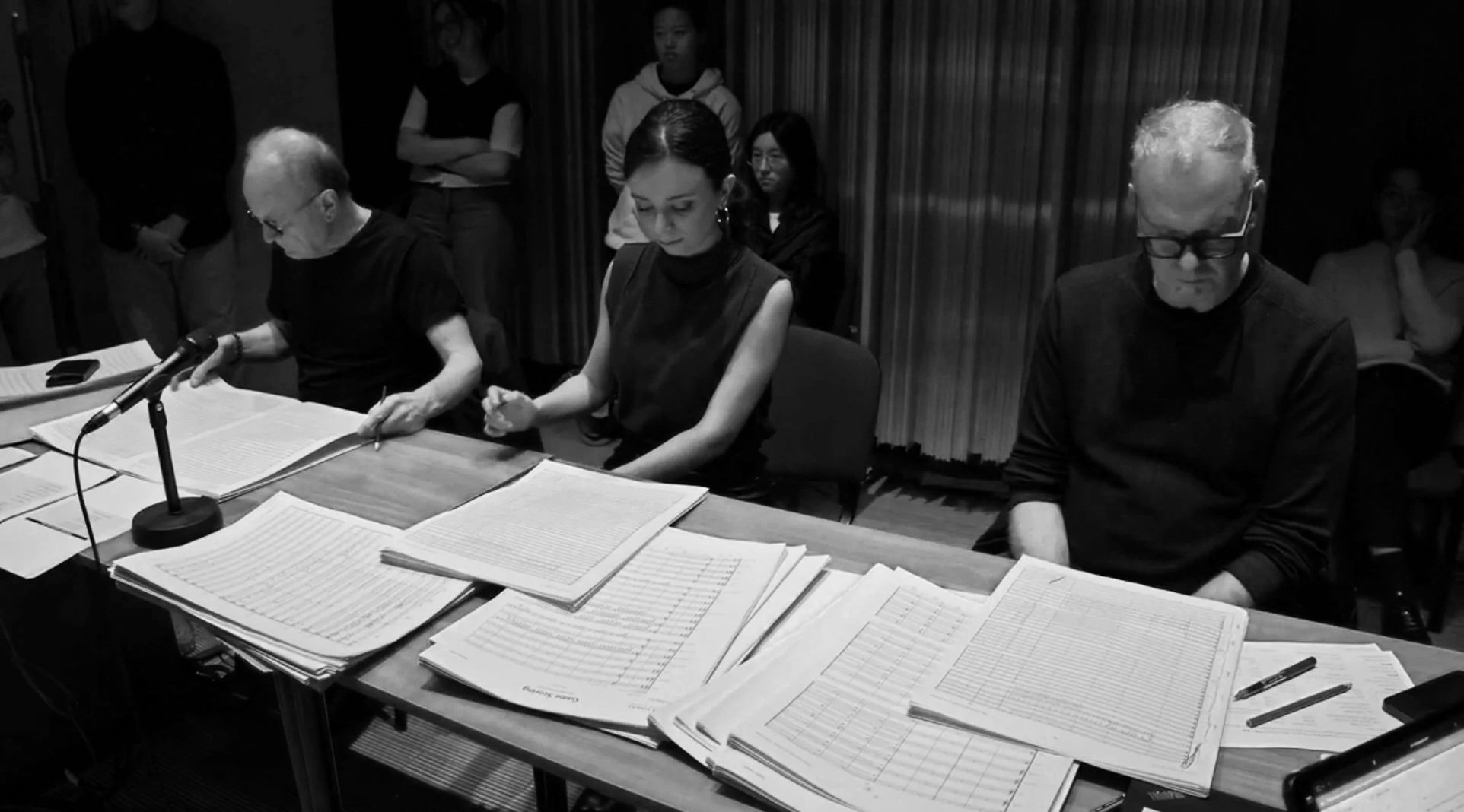 Three people sitting at a table with multiple stacks of papers in front of them. The person on the left is reading and writing, the woman in the middle has her hands clasped, and the man on the right has his head bowed, all in a room with others stan