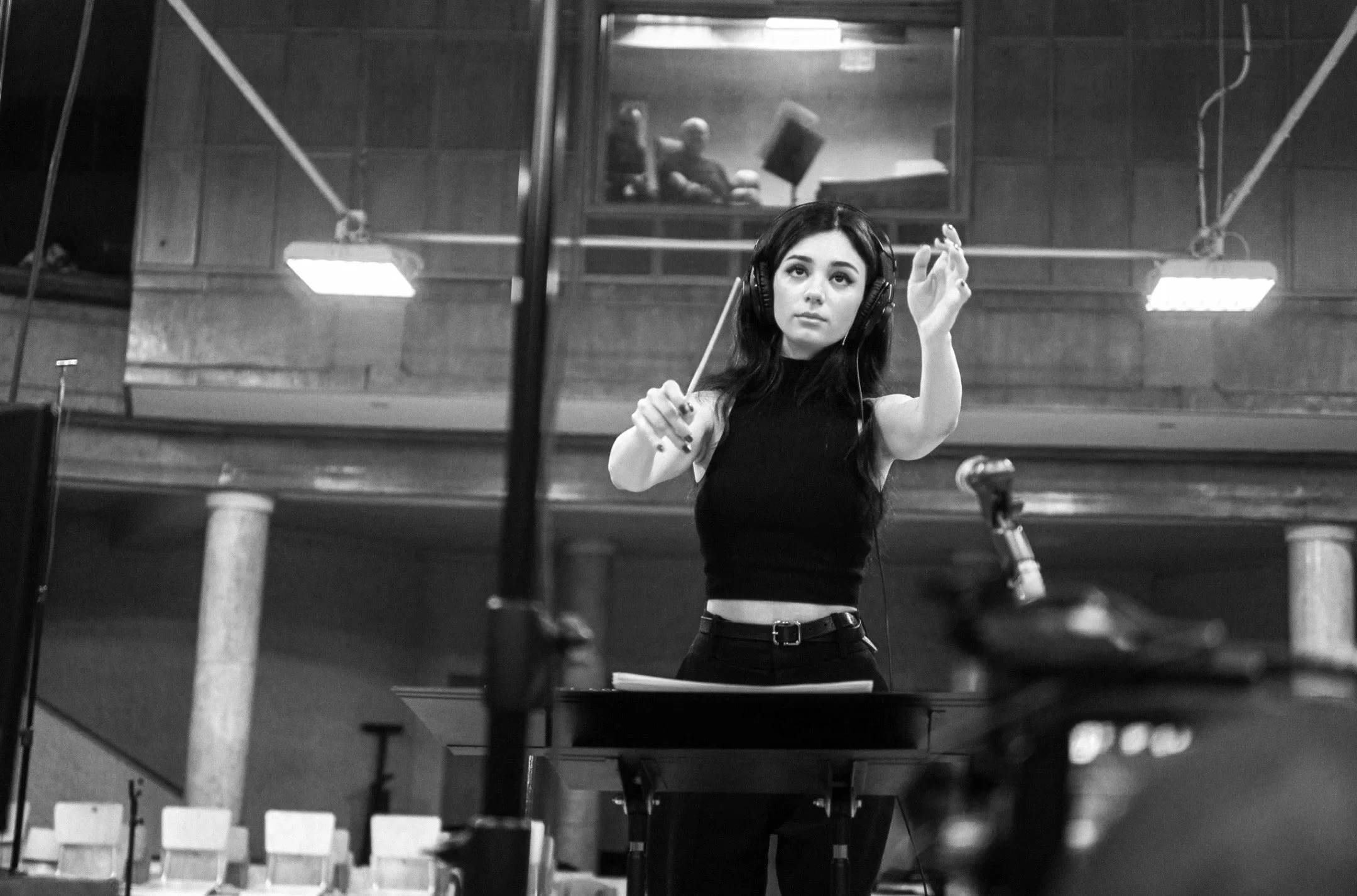 A young woman with long dark hair, wearing a black sleeveless crop top and headphones, conducts an orchestra with a baton in her right hand inside a concert hall.