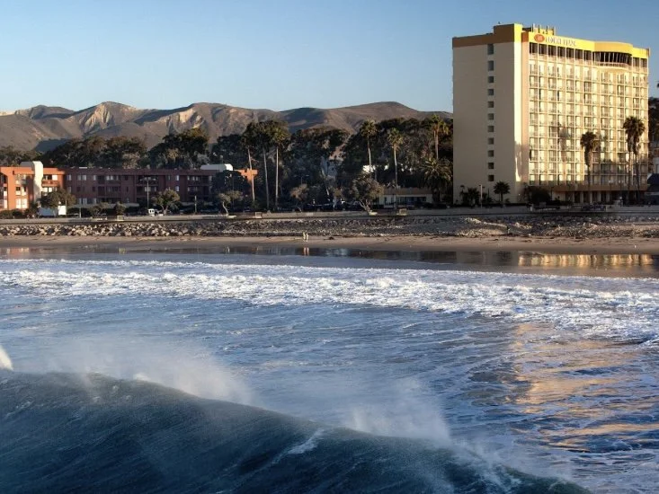 Waves crashing near the shoreline with buildings, palm trees, and mountains in the background during sunset.