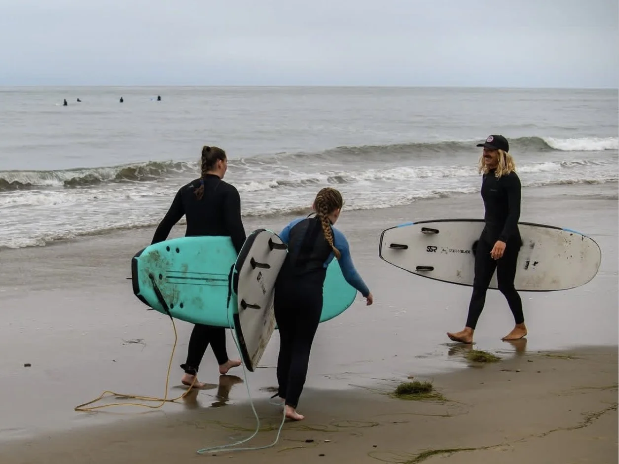 Three women walking on the beach with surfboards, near the ocean with waves and a cloudy sky.