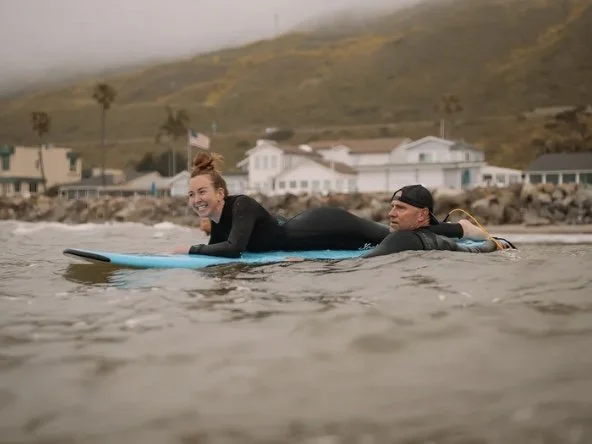 A woman and a man on a surfboard in the water near a beach with houses and hills in the background.