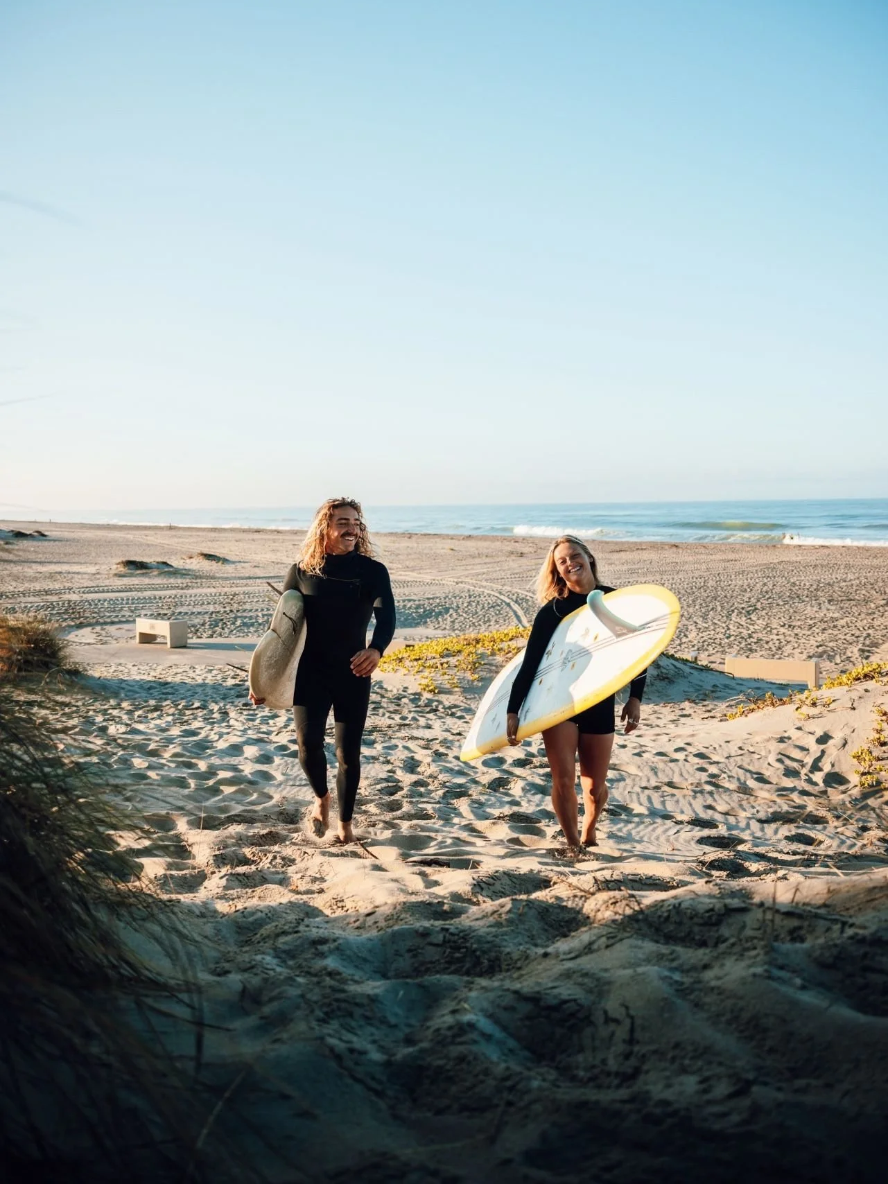 Two women at the beach, one holding a surfboard, walking on the sand towards the ocean.