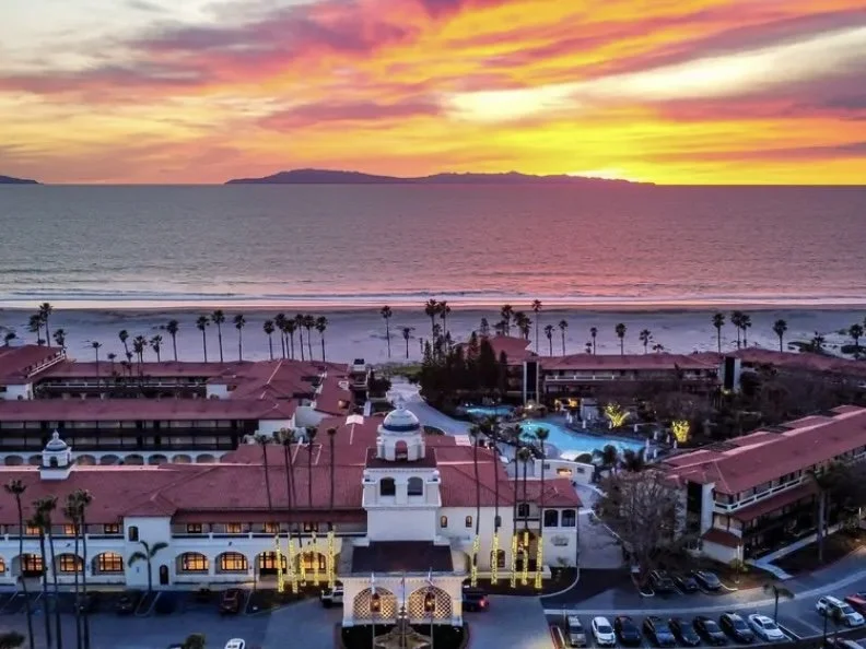 Sunset view of a beach resort with palm trees, a pool, and the ocean in the background.