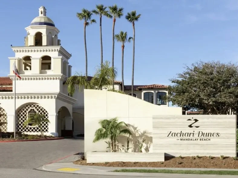 Entrance sign for Zachari Dunes on Mandalay Beach with white building, palm trees, and clear blue sky in the background.