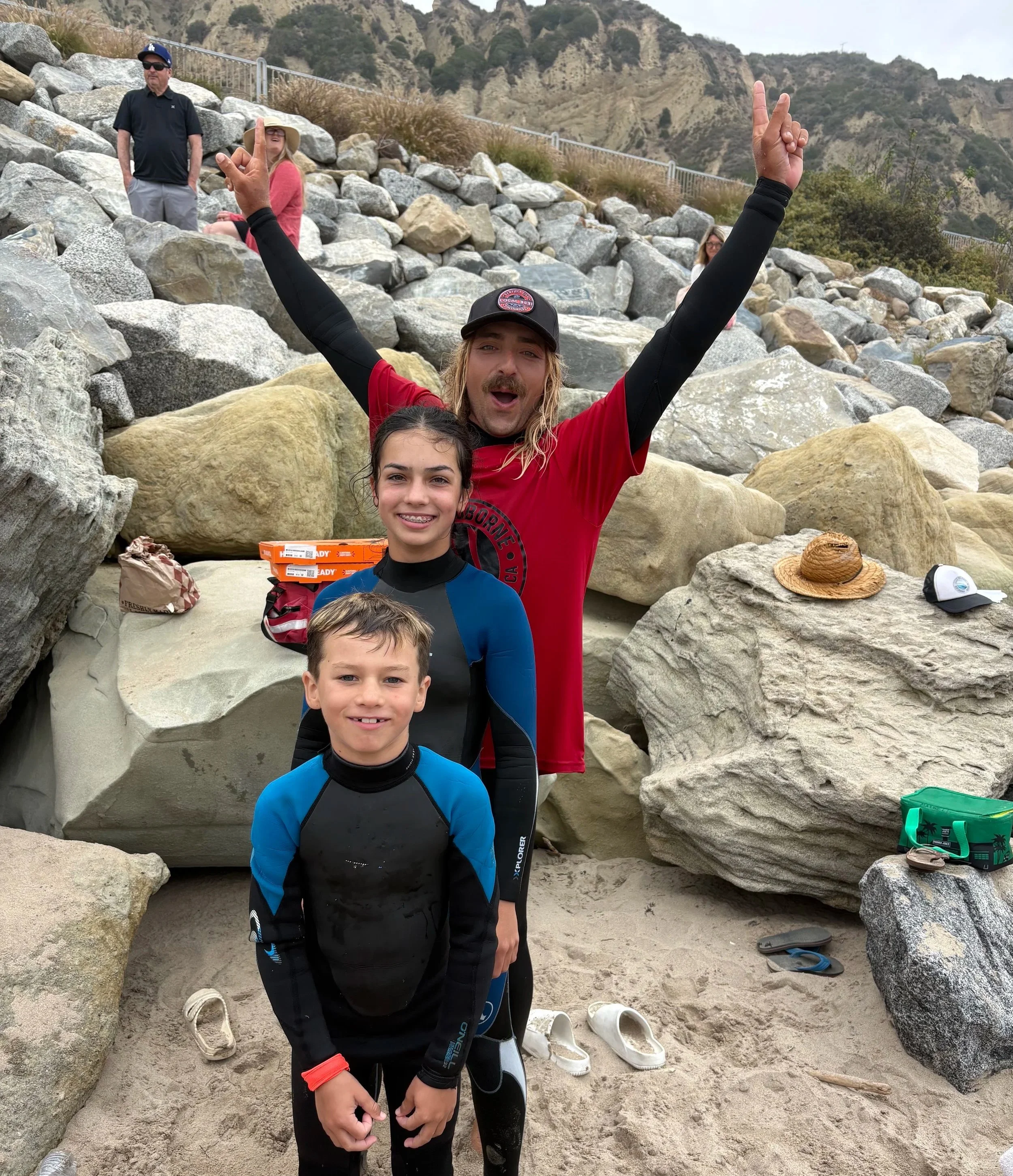 A group of five people on a rocky beach, with mountains in the background. A man with long blond hair, a beard, and a black cap is raising his arms in the air, making peace signs. Two boys are standing in front, wearing wetsuits, with one holding his hands together. Two women are sitting on rocks, one wearing a wide-brimmed hat and pink clothing, and the other wearing glasses. Items like shoes, a straw hat, and bags are scattered on the sand.