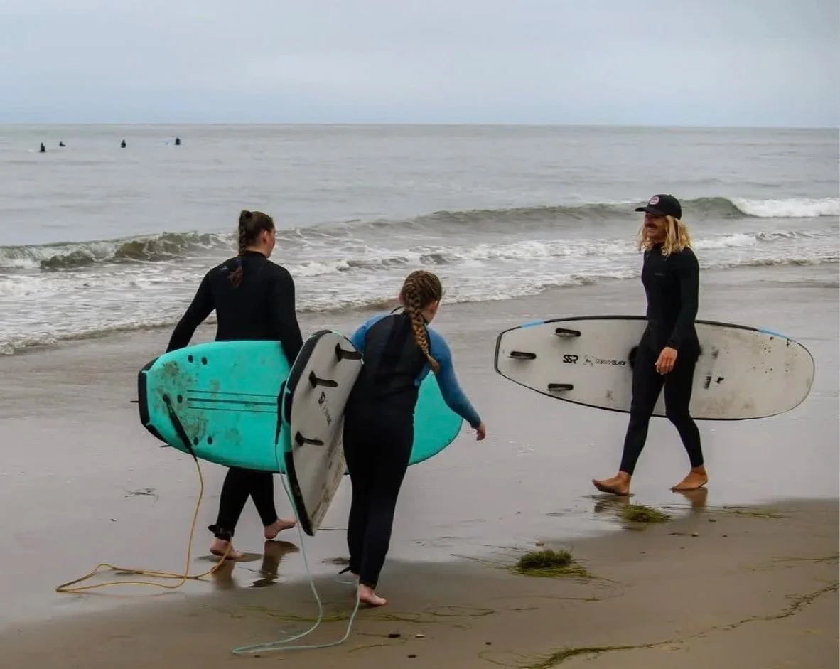 Three women walking on the beach holding surfboards, with waves in the background and others surfing further out in the water.