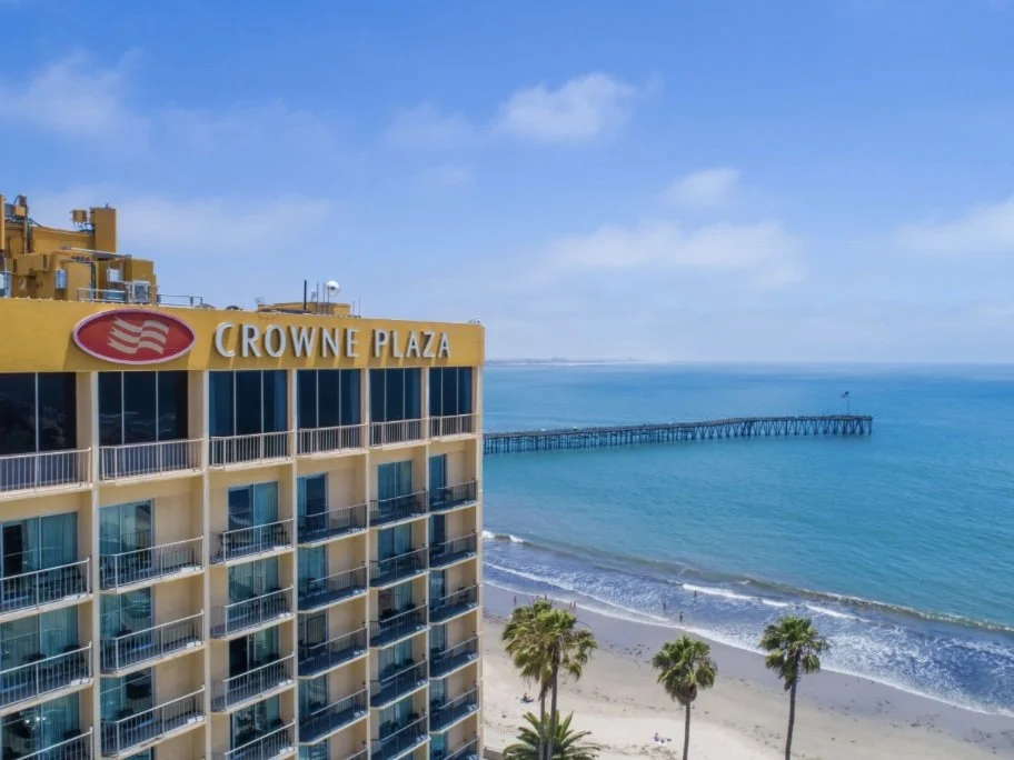 View of the Crowne Plaza hotel on the beach with a pier extending into the ocean, blue sky, and palm trees.