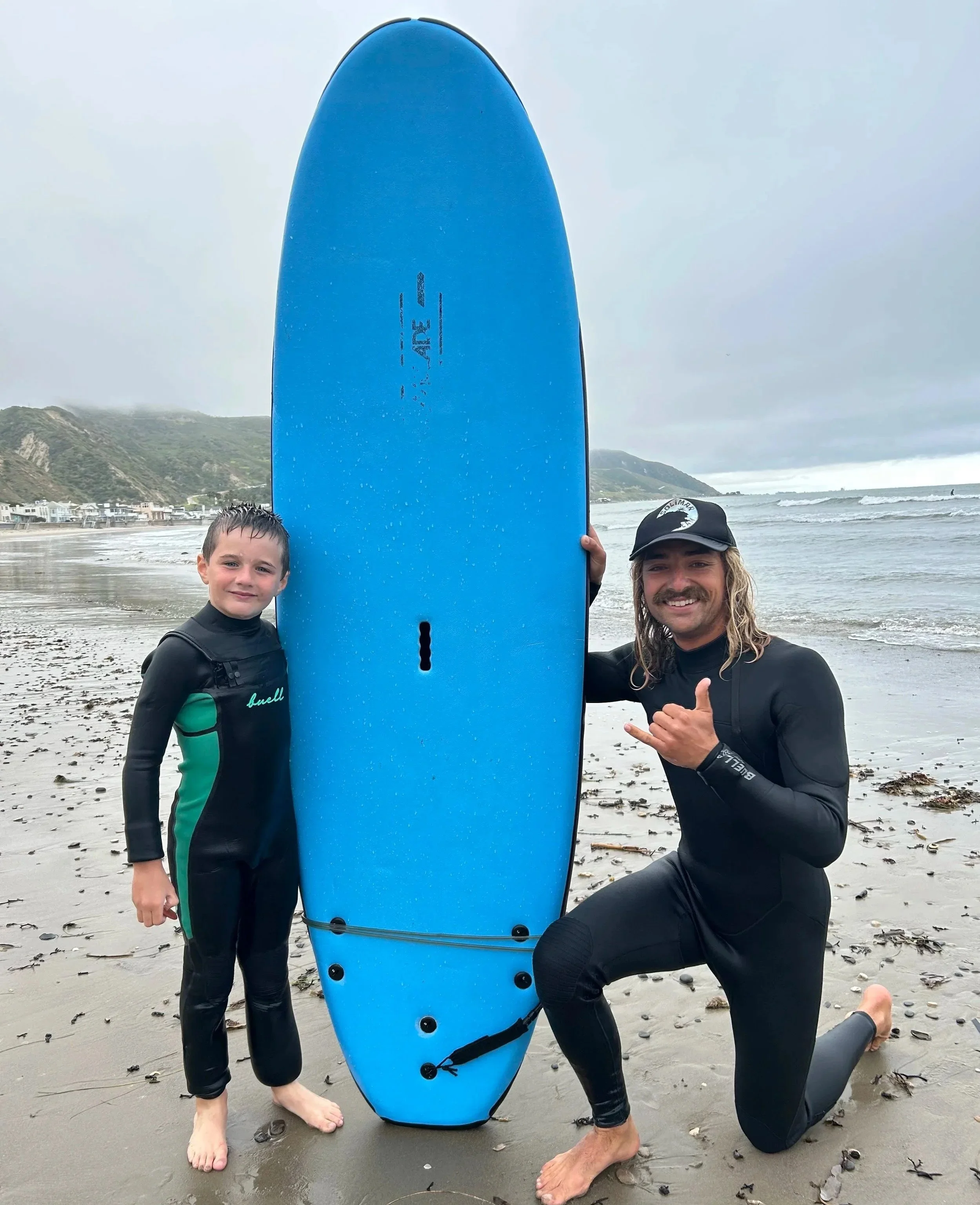 Two people at the beach, one young boy and one adult male, posing with a large blue surfboard. The boy is standing, the man is kneeling on the sand, giving a thumbs-up and making a shaka sign. Overcast sky, shoreline with water, hills, and houses in the background.