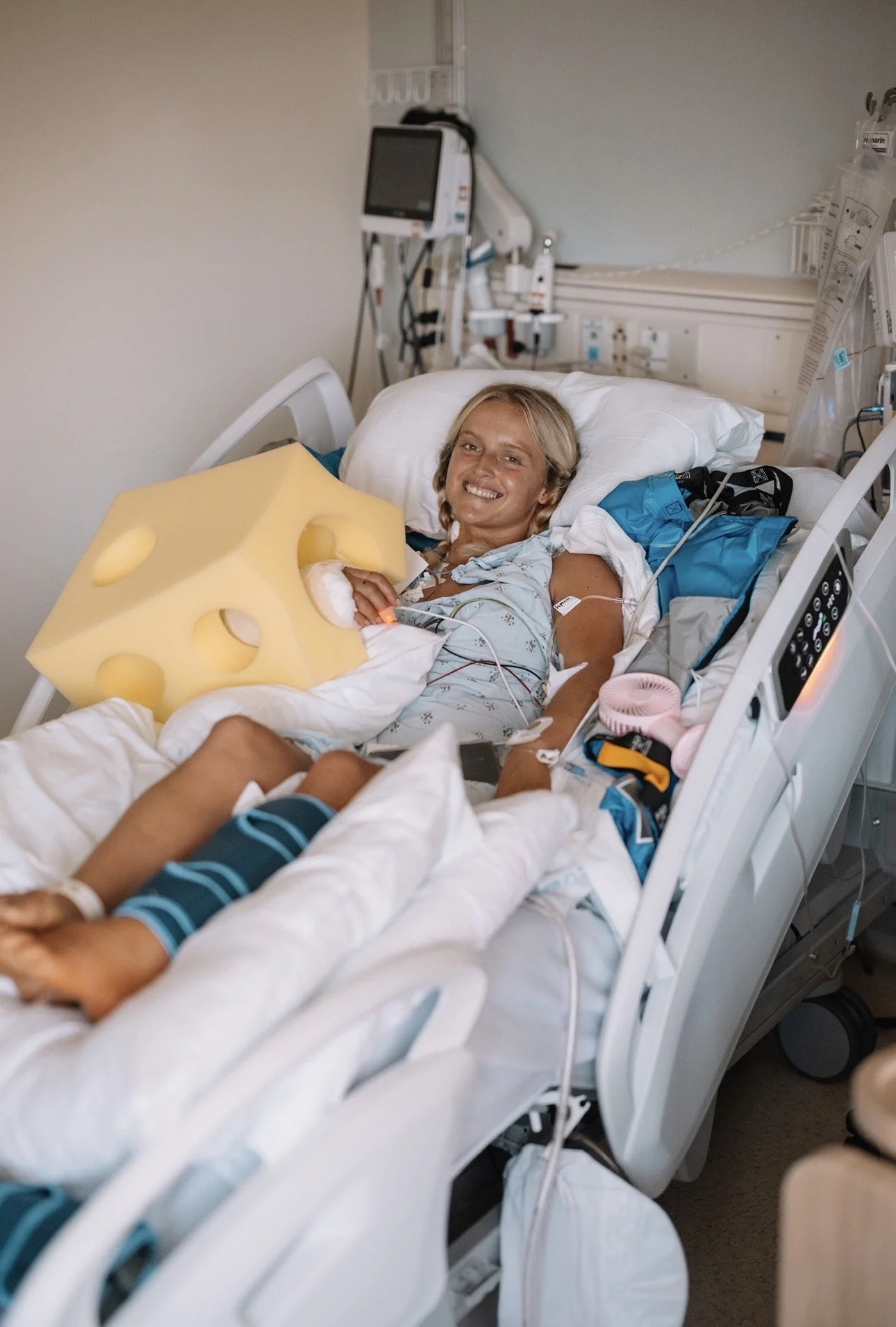 A woman lying in a hospital bed smiling, holding a large yellow foam cheese block, surrounded by medical equipment and wires in a hospital room.