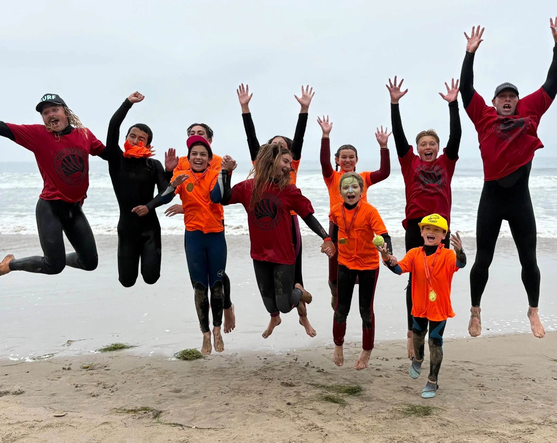 Children and adults jumping and playing on a beach with the ocean in the background, wearing colorful wetsuits and rash guards, enjoying a fun day at the seaside.