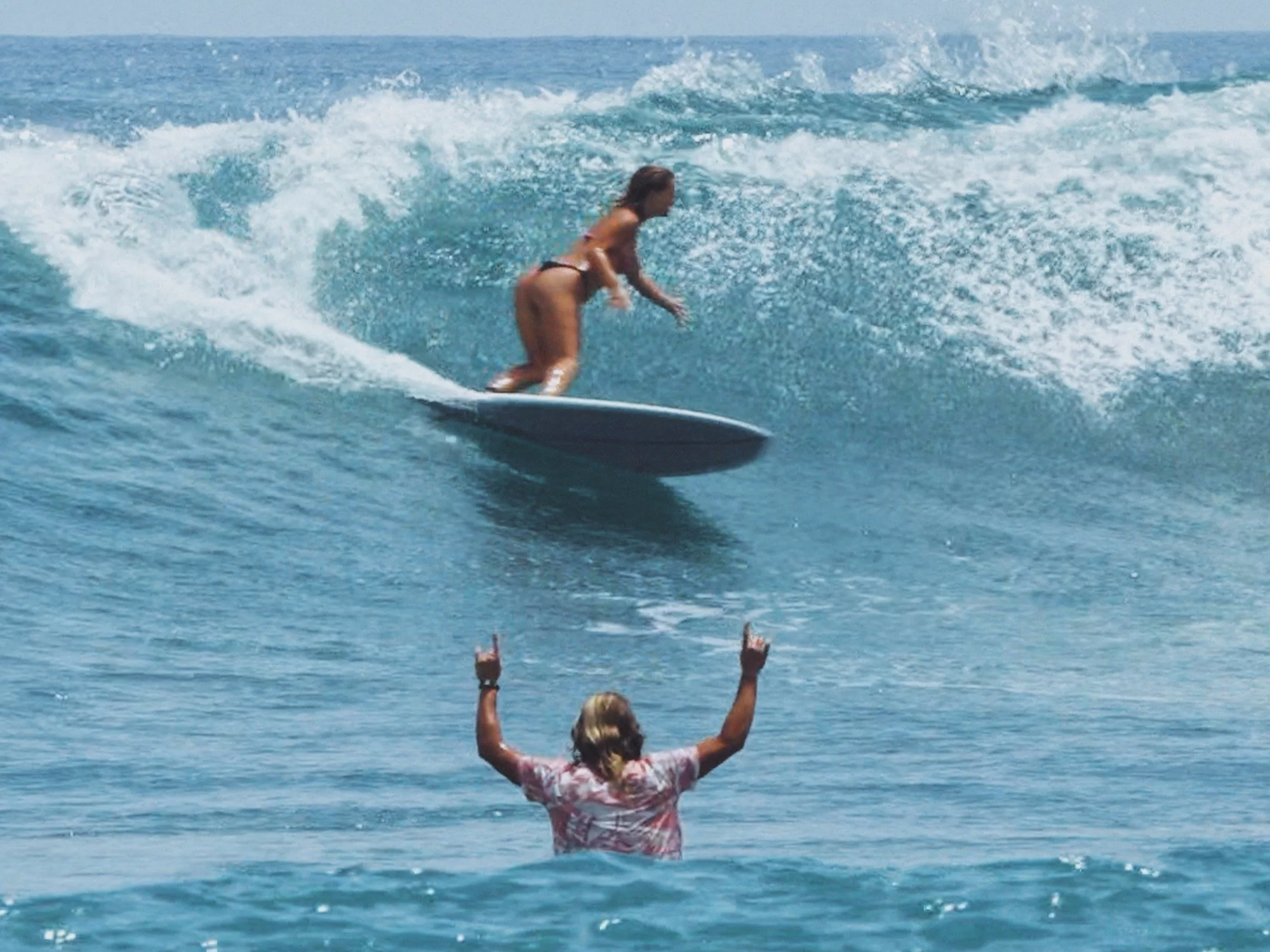 A woman with blonde hair wearing a pink and white dress standing in the water with her arms raised, watching as a person on a surfboard rides a wave in the ocean.